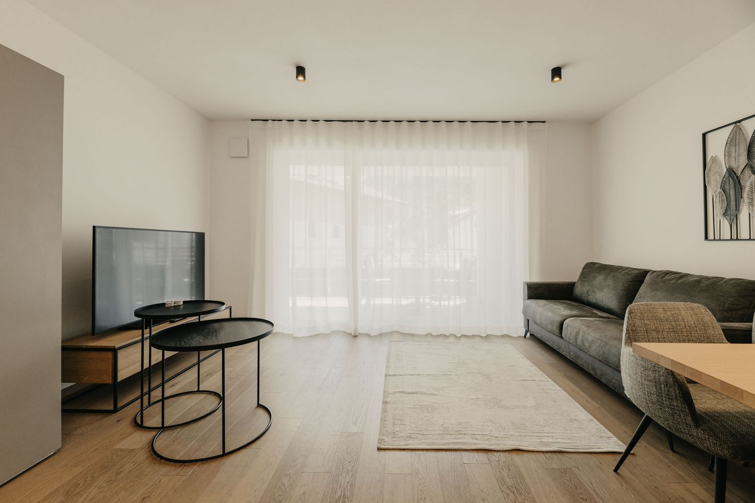 Ein minimalistisch eingerichtetes Wohnzimmer mit einem grauen Sofa, zwei schwarzen Beistelltischen, einem Fernseher auf einem Holz-Sideboard und einem hellen Teppich auf hellem Holzboden.