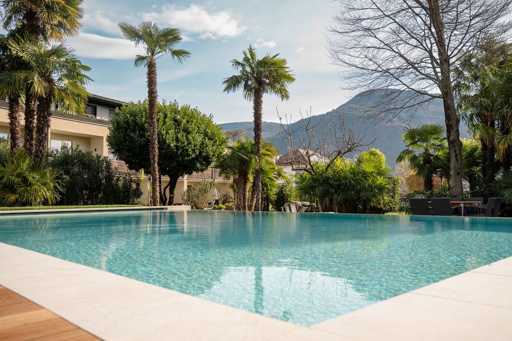 Vista su una piscina all'aperto dall'acqua cristallina con palme circostanti, un edificio alberghiero e montagne sullo sfondo in una giornata di sole.