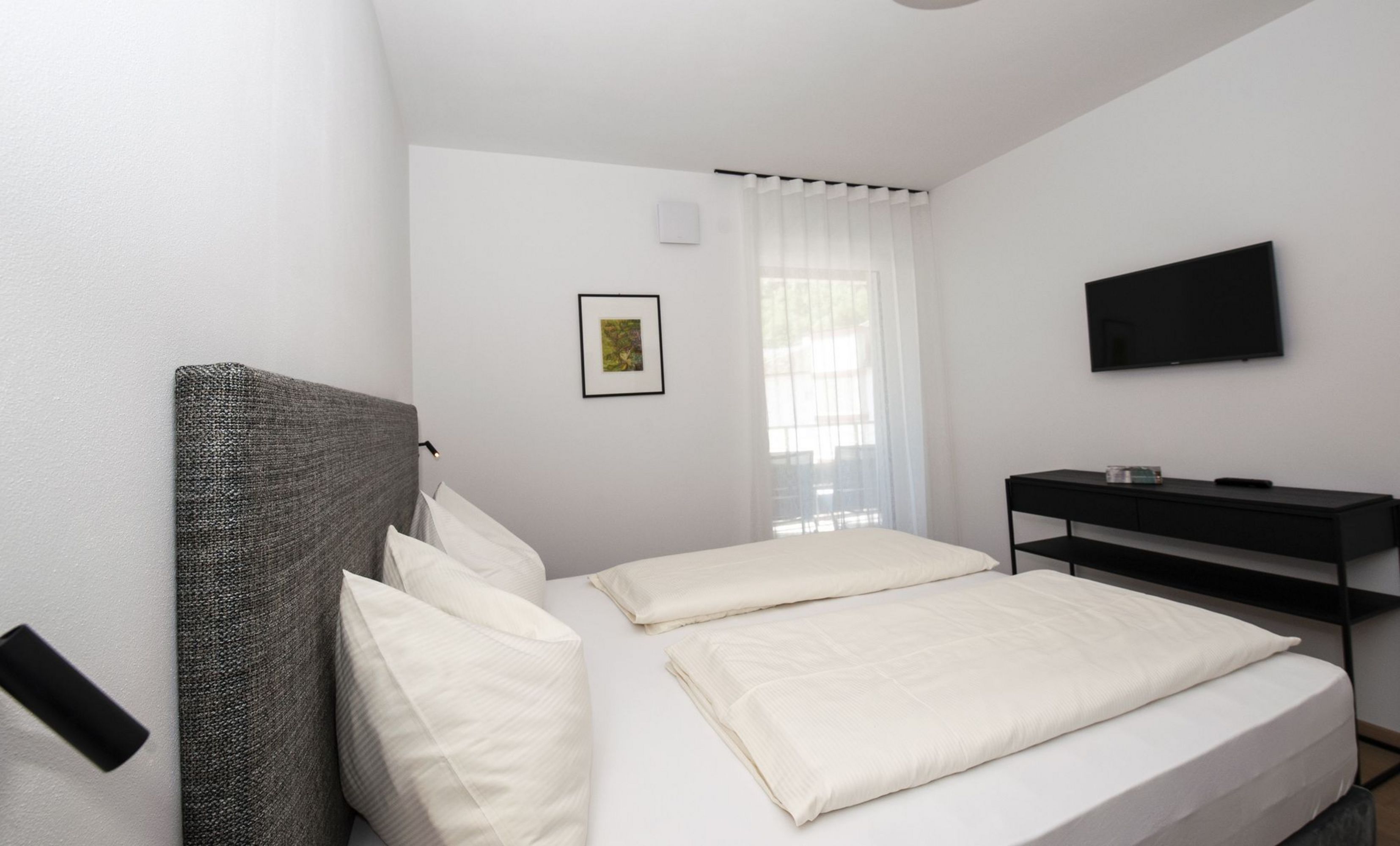 Ein minimalistisch eingerichtetes Hotelzimmer mit einem Doppelbett, weißer Bettwäsche, einem grauen Kopfteil, einem Fernseher an der Wand und einem kleinen schwarzen Konsolentisch vor einem Fenster mit halbtransparentem Vorhang.
