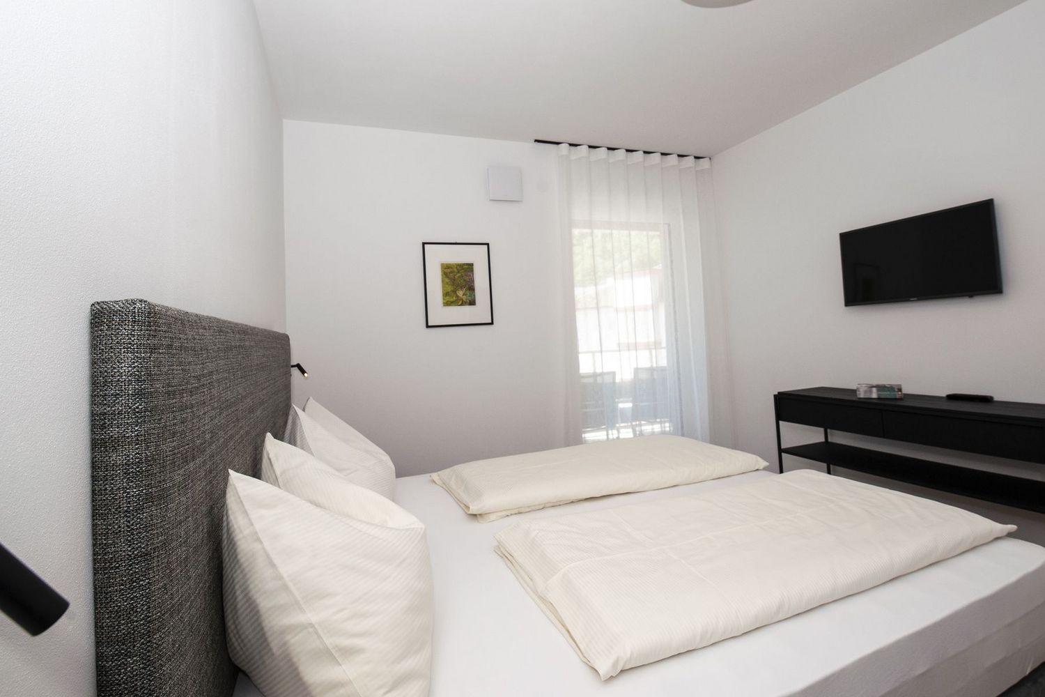 Ein minimalistisch eingerichtetes Hotelzimmer mit einem Doppelbett, weißer Bettwäsche, einem grauen Kopfteil, einem Fernseher an der Wand und einem kleinen schwarzen Konsolentisch vor einem Fenster mit halbtransparentem Vorhang.