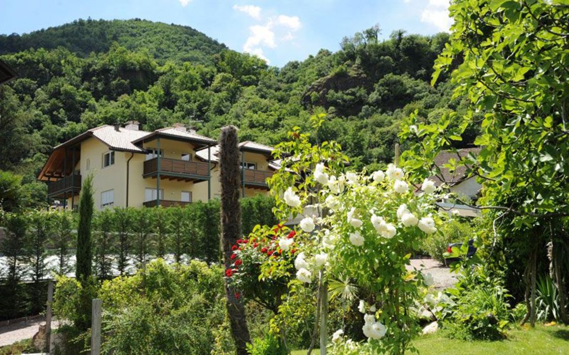 Ein gelbes Mehrfamilienhaus mit braunen Balkonen steht vor einem bewaldeten Berg, im Vordergrund blühen weiße und rote Rosen in einem Garten.