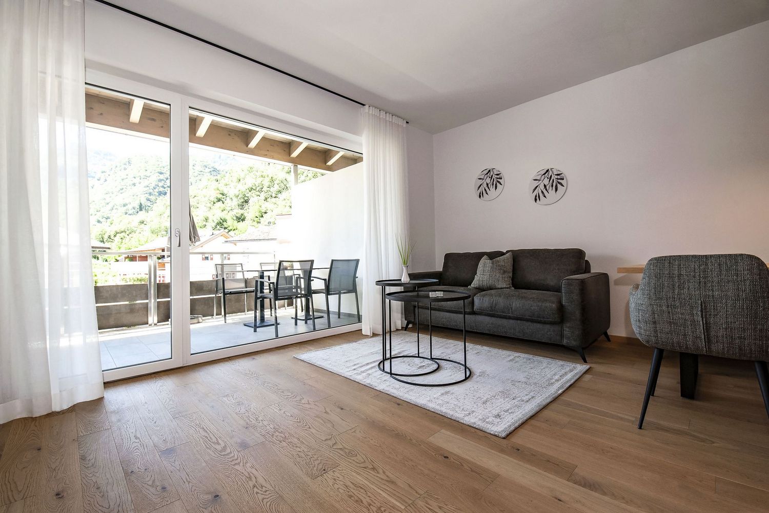Ein lichtdurchflutetes Wohnzimmer mit grauem Sofa, modernen Couchtischen und Holzboden, das durch eine große Glasfront mit einem Balkon verbunden ist.