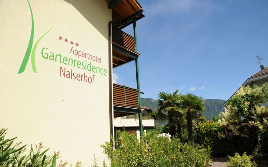 Seitenansicht der hellen Außenfassade des Apparthotel Gartenresidence Nalserhof mit Balkonen, Palmen und grüner Vegetation vor einem blauen Himmel.