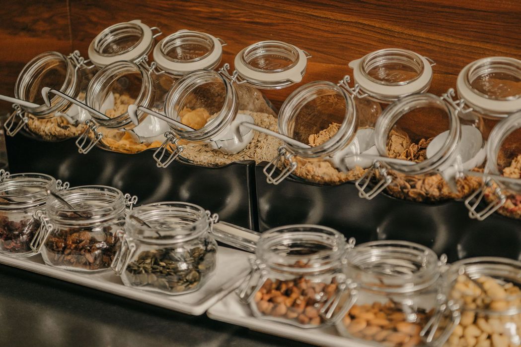 Eine Auswahl verschiedener Cerealien und Nüsse in Glasgefäßen, die in einem Frühstücksbuffet auf einer dunklen Ablage präsentiert werden.