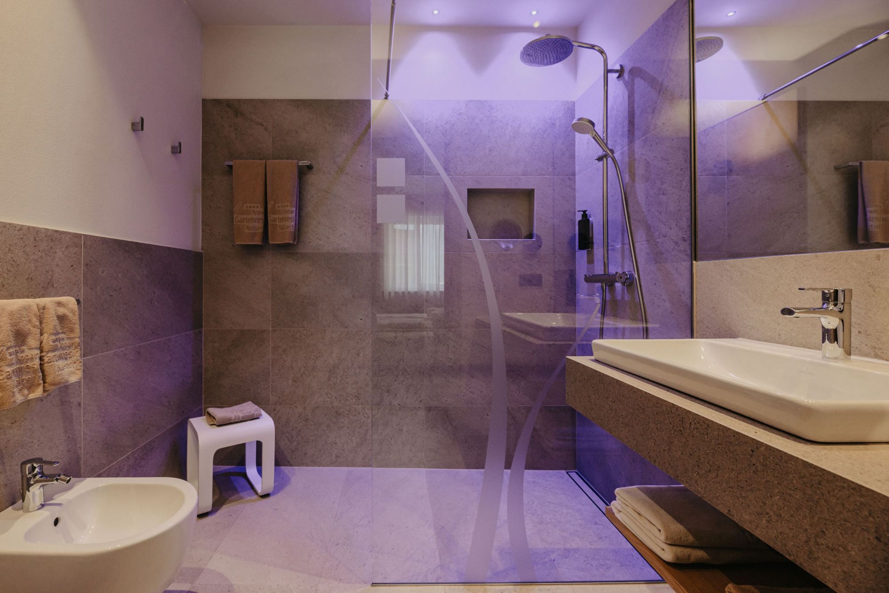 Modernes Badezimmer mit Dusche mit Glastrennwand, Waschbecken, Bidet und grauen Fliesen, beleuchtet mit violettem Licht.