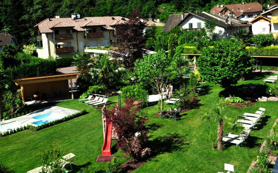 Ein von oben fotografierter Gartenbereich eines Hotels mit Liegestühlen, einem kleinen Pool, einer Kinderrutsche und vielen Bäumen vor mehreren Häusern.