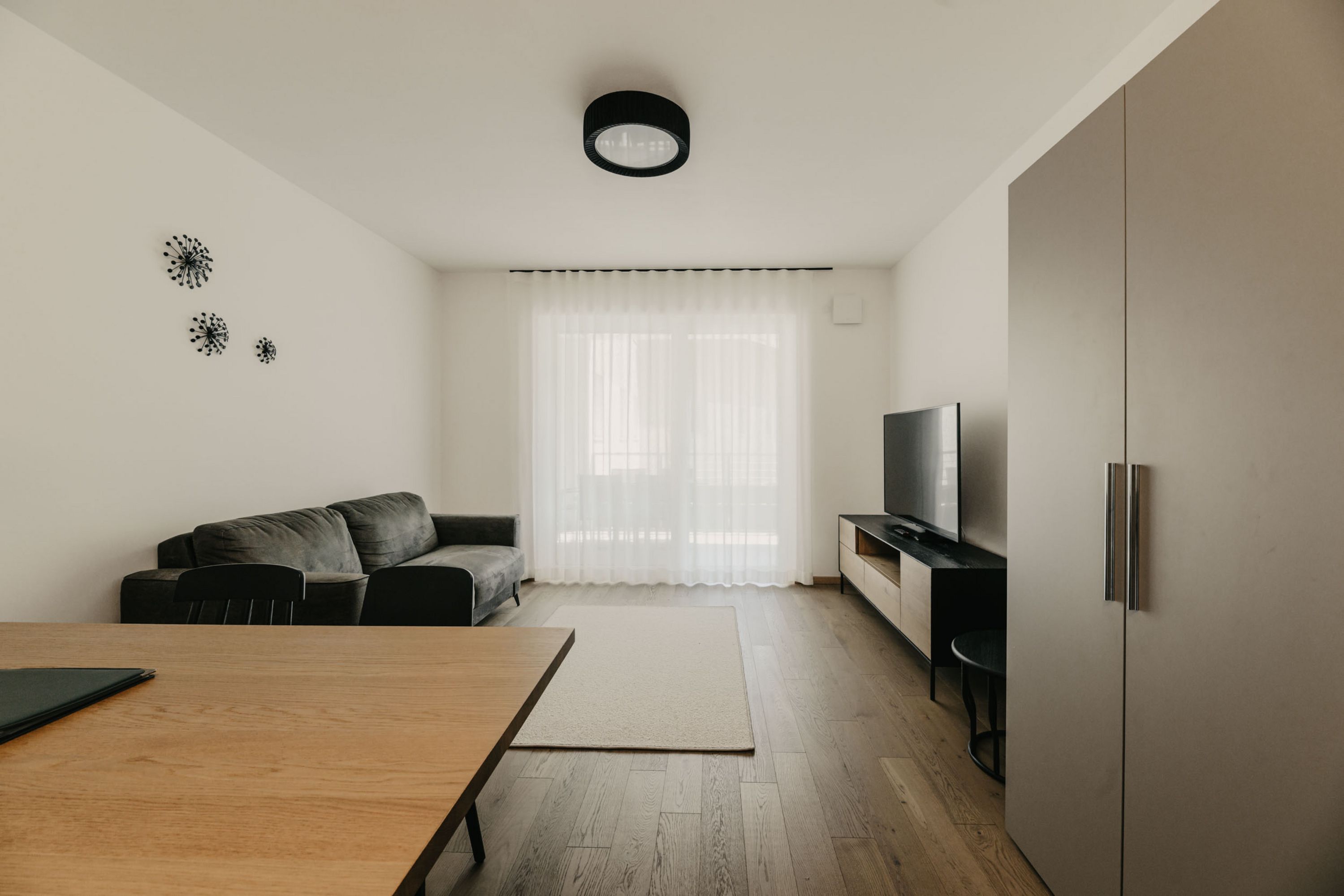 Ein minimalistisches Wohnzimmer mit einem dunkelgrauen Sofa, einem Holztisch im Vordergrund, einem TV-Board und einer hellen Fensterfront mit Vorhängen.