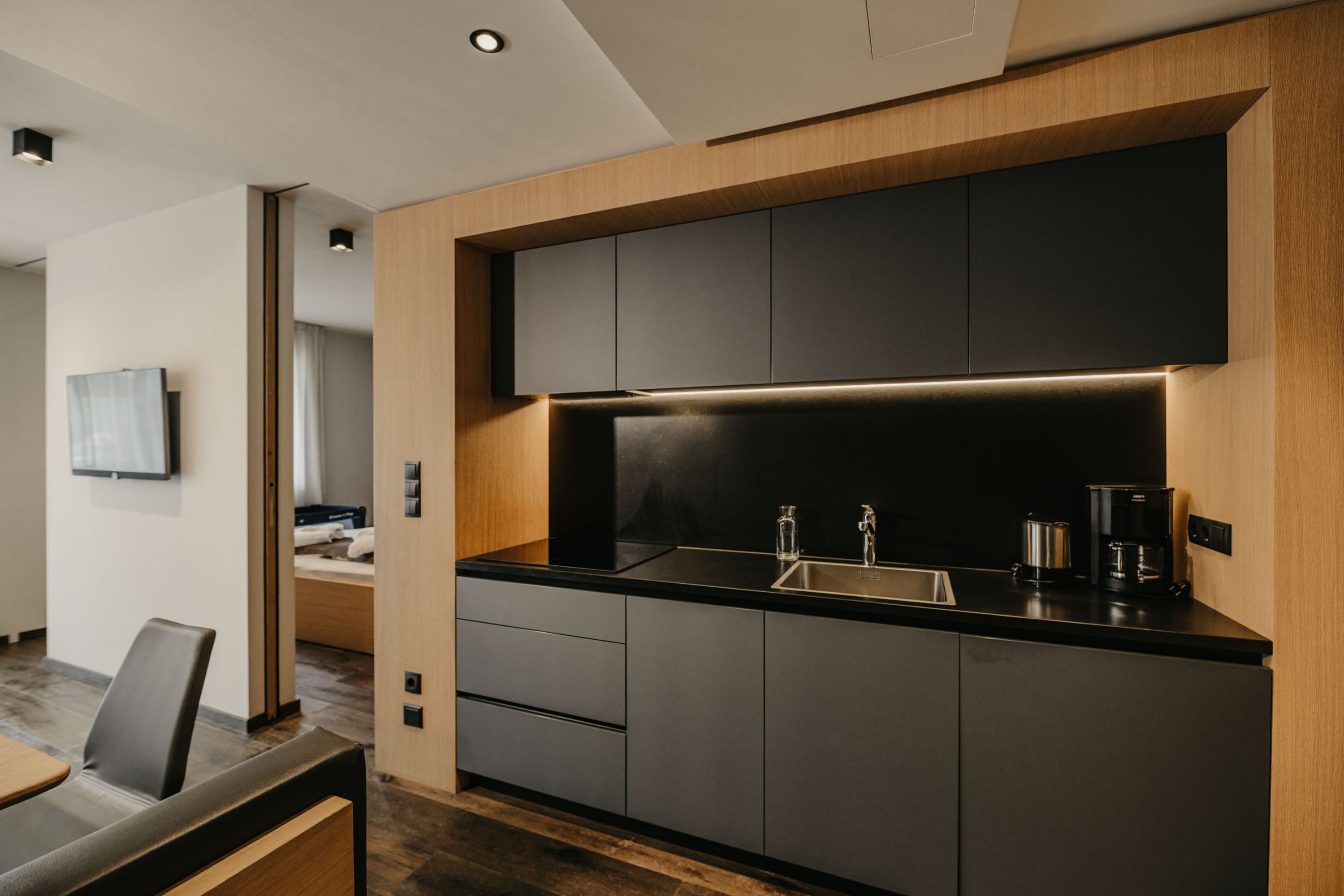 Eine moderne Küchenzeile mit mattschwarzen Schränken, einer dunklen Arbeitsplatte und hellem Holzrahmen, daneben ein Durchgang zu einem Hotelzimmer.