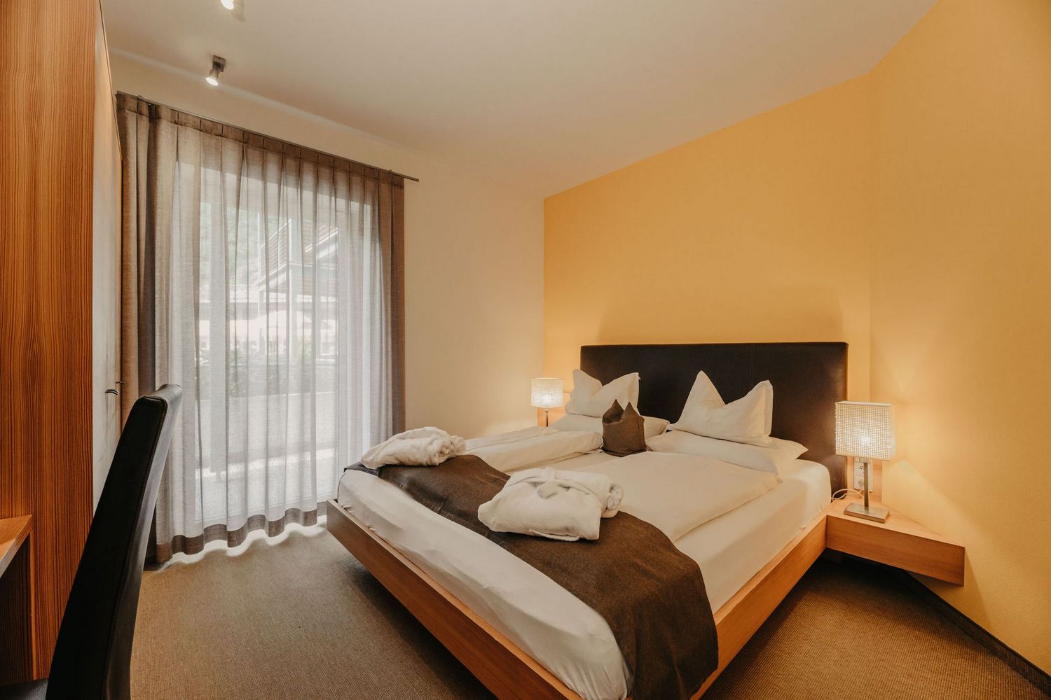 Ein gemütliches Hotelzimmer mit einem Doppelbett, einem Holzbettrahmen, weißen Kissen und zwei Bademänteln, flankiert von Nachttischlampen und einem Fenster mit Vorhang.