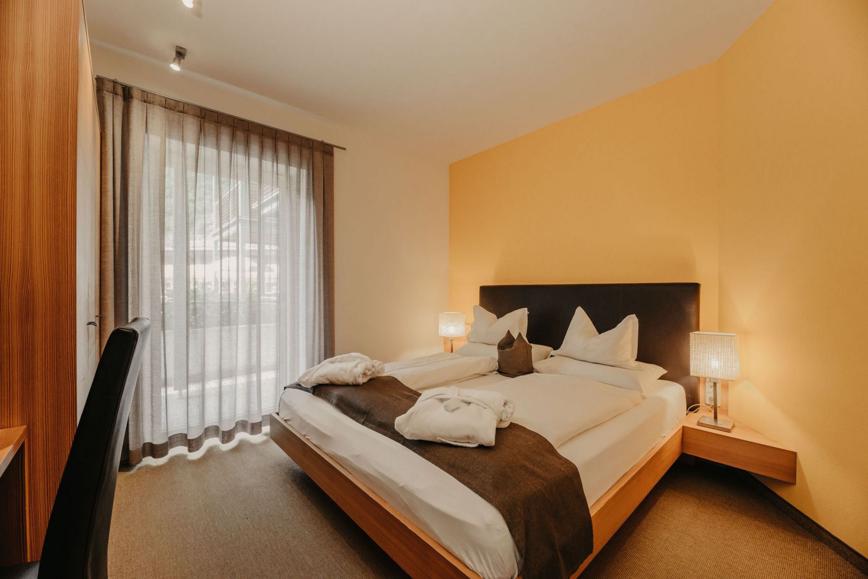 Ein gemütliches Hotelzimmer mit einem Doppelbett, einem Holzbettrahmen, weißen Kissen und zwei Bademänteln, flankiert von Nachttischlampen und einem Fenster mit Vorhang.