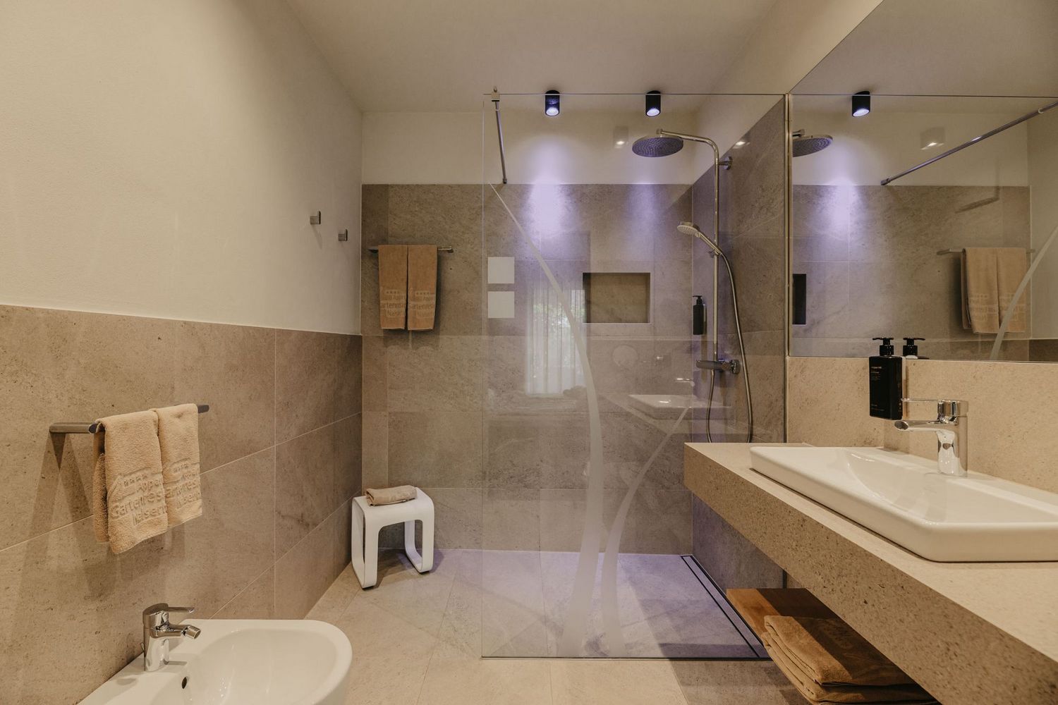 Ein modernes Badezimmer mit einer bodenebenen Glasdusche, einem Waschbecken auf einem Steinsockel, einem Bidet und beige gefliesten Wänden.