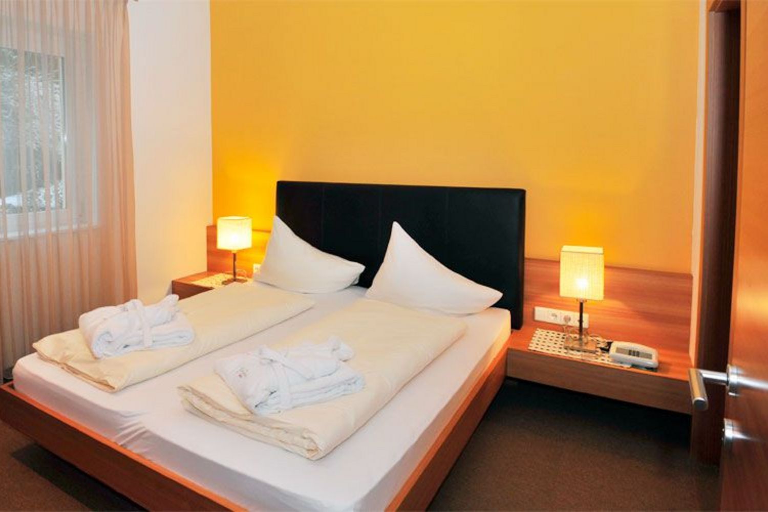 Ein Hotelzimmer mit einem Doppelbett, zwei Nachttischlampen und zwei auf den Betten liegenden Bademänteln vor einer gelben Wand.