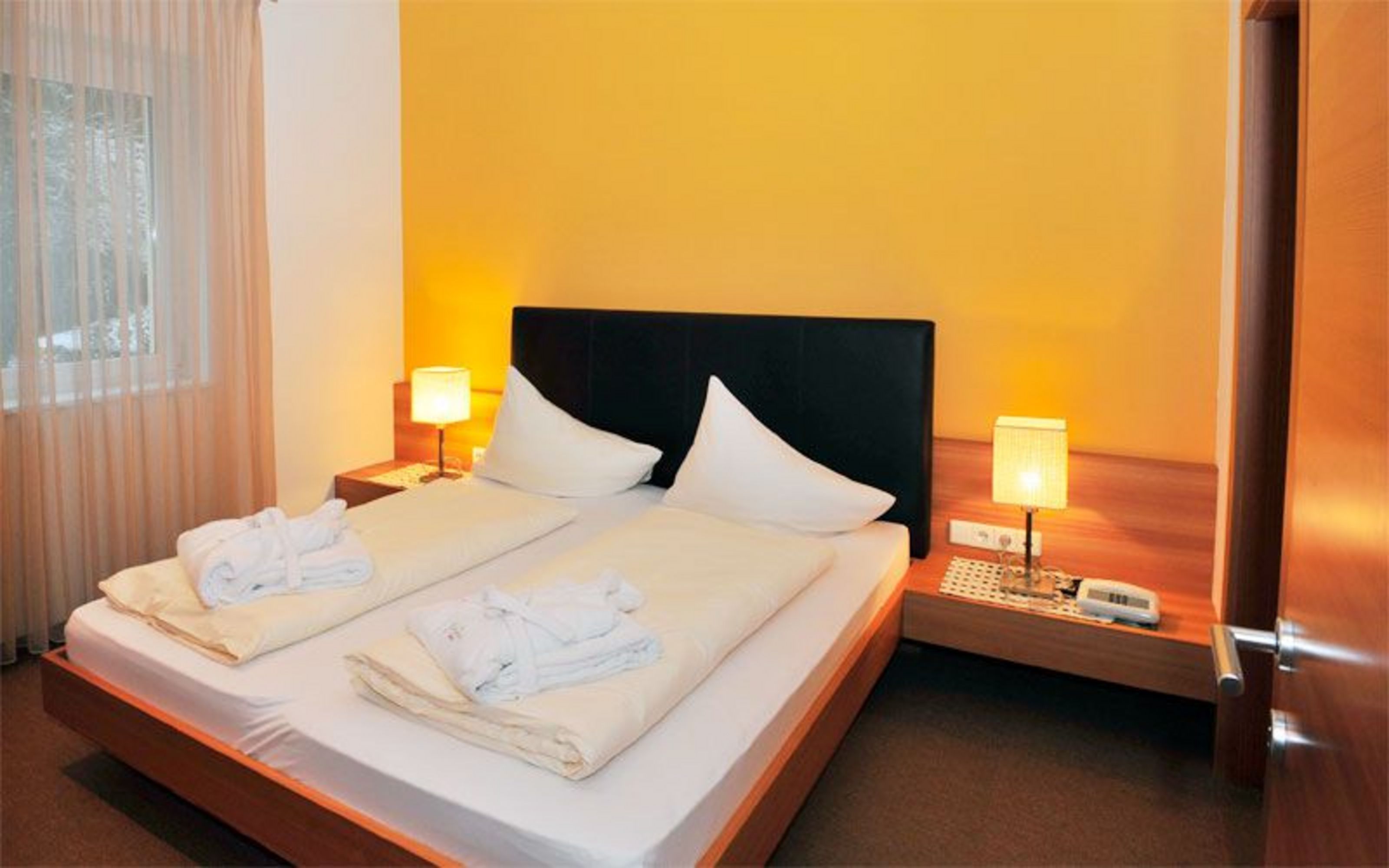 Ein Hotelzimmer mit einem Doppelbett, zwei Nachttischlampen und zwei auf den Betten liegenden Bademänteln vor einer gelben Wand.