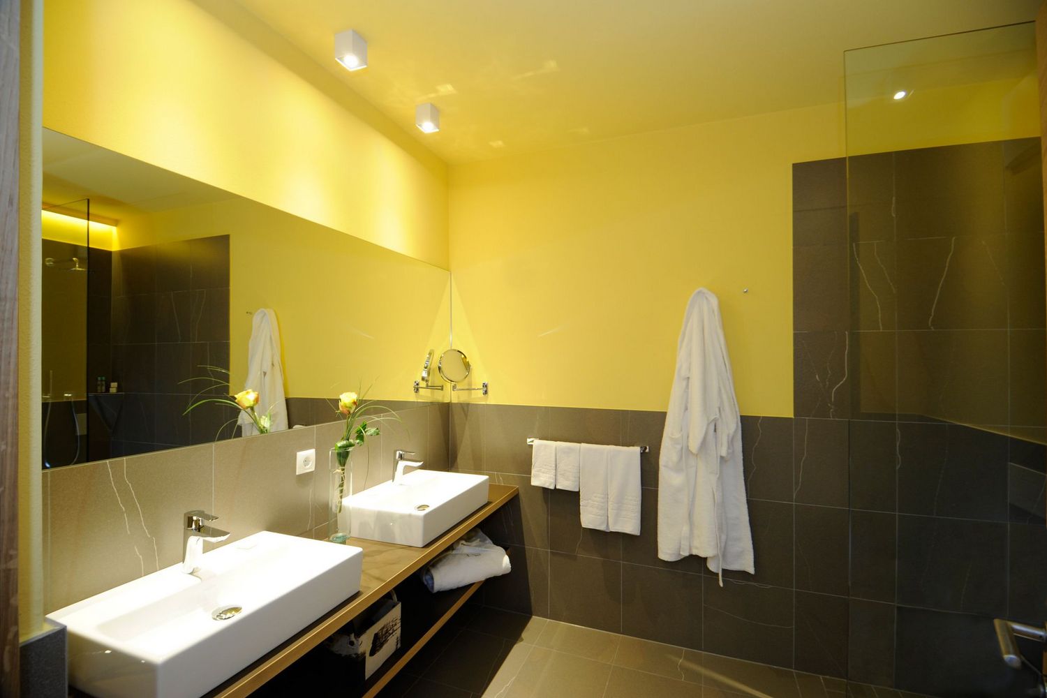 Modernes Badezimmer mit zwei weißen Waschbecken, grauen Fliesen, leuchtend gelben Wänden und weißen Bademänteln.