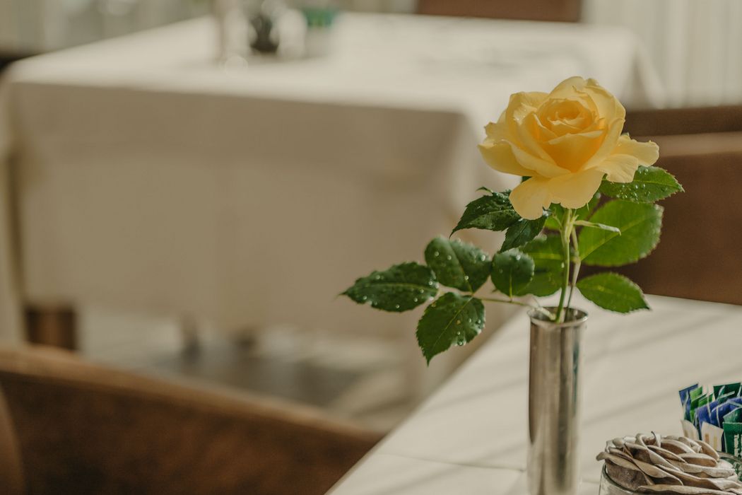 Eine einzelne gelbe Rose in einer schmalen Metallvase steht auf einem Tisch in einem Restaurant.