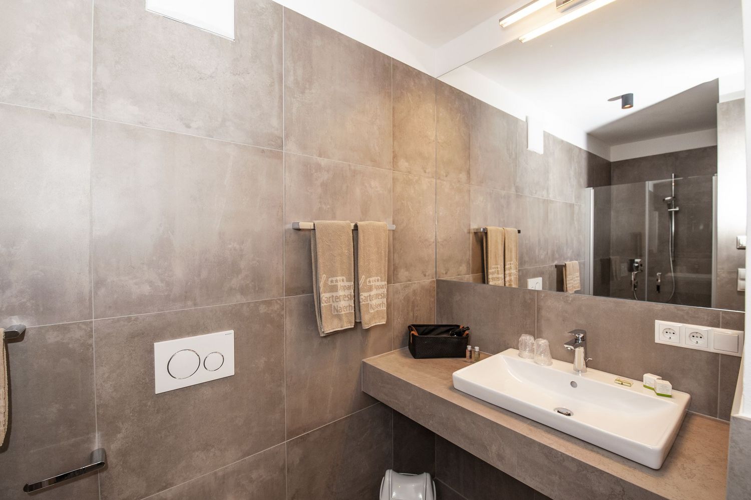 Ein modernes Badezimmer mit grauen Fliesen, einem weißen Waschbecken auf einem Waschtisch und einem großen Wandspiegel.