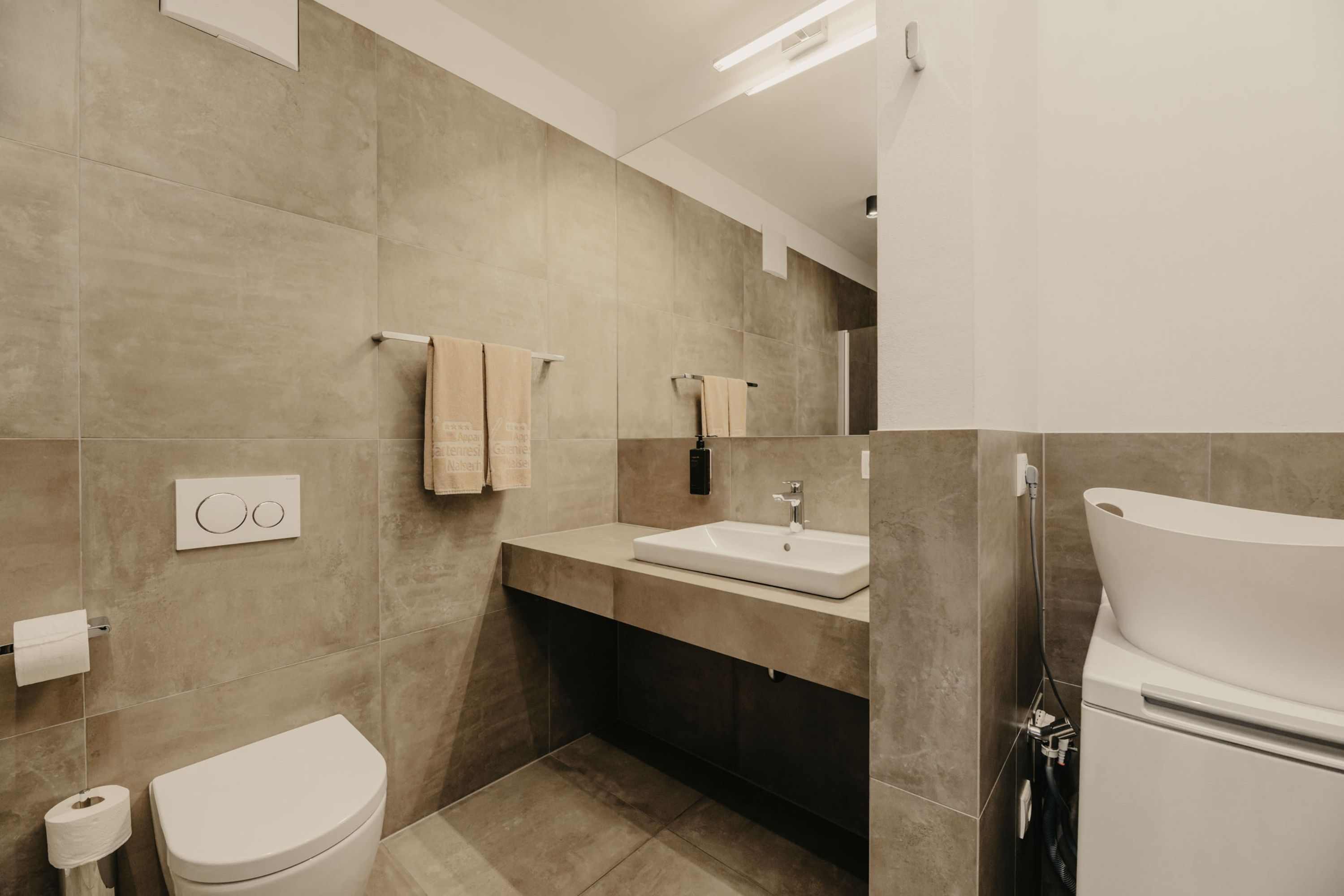 Ein modernes Badezimmer mit grauen Fliesen, einem weißen Waschbecken auf einem wandhängenden Waschtisch, einer Toilette und zwei beigen Handtüchern an einem Handtuchhalter.