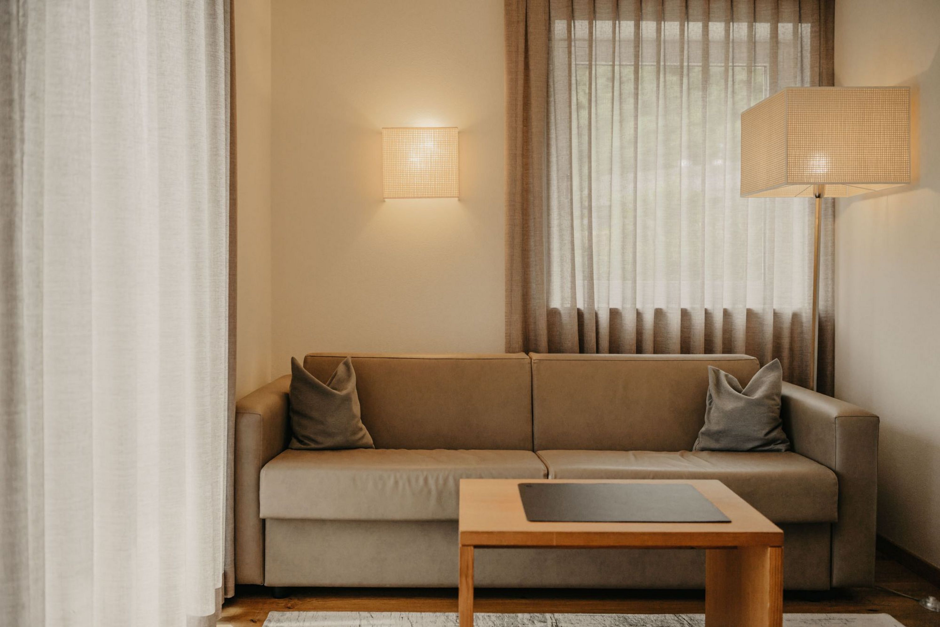 Ein beige Sofa mit zwei grauen Kissen steht in einem modern und gemütlich eingerichteten Wohnbereich vor einem Fenster mit beigen Vorhängen, ergänzt durch eine Stehlampe und eine Wandleuchte.
