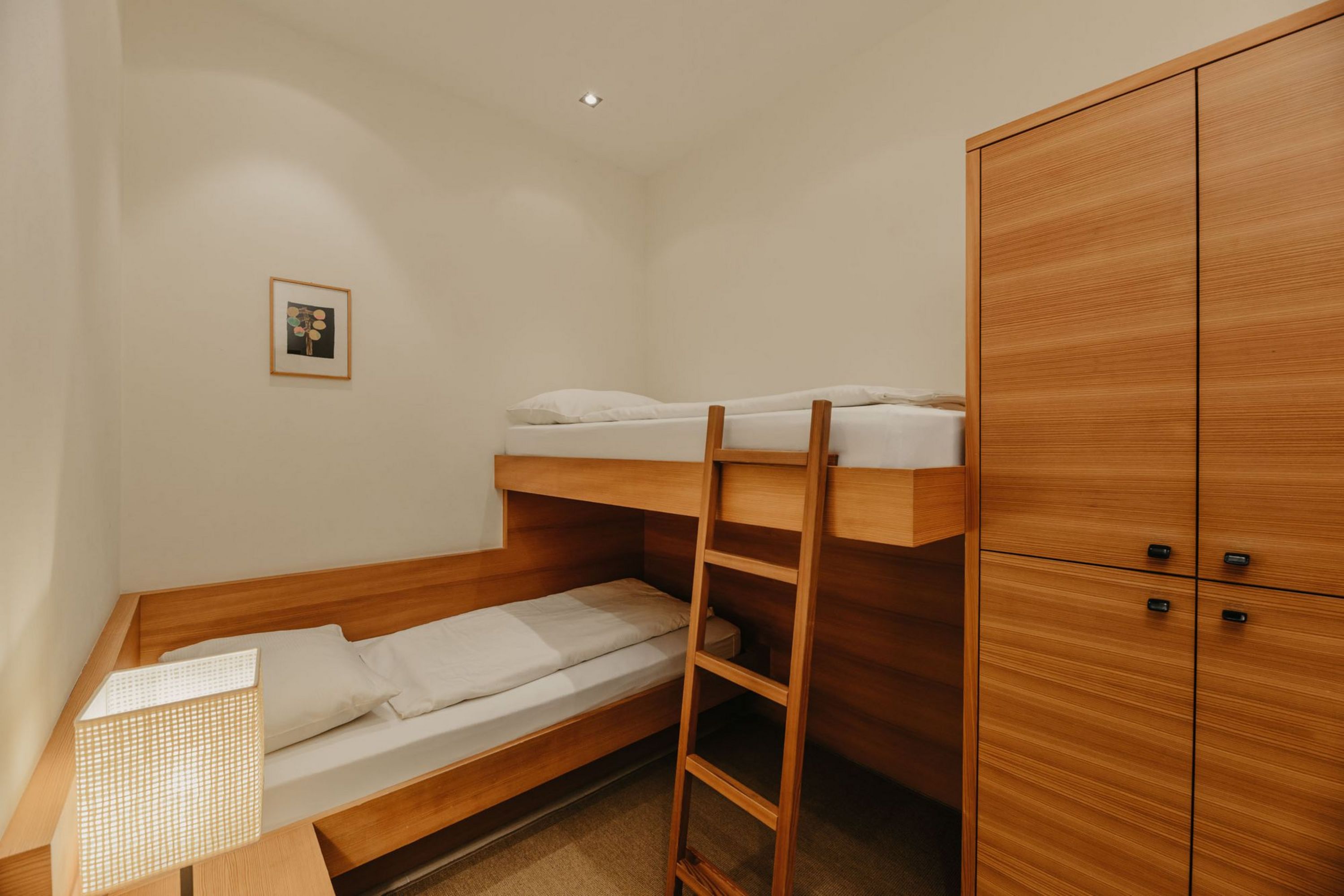 Ein kleines Hotelzimmer mit einem hölzernen Etagenbett, einem Kleiderschrank und einer Nachttischlampe.