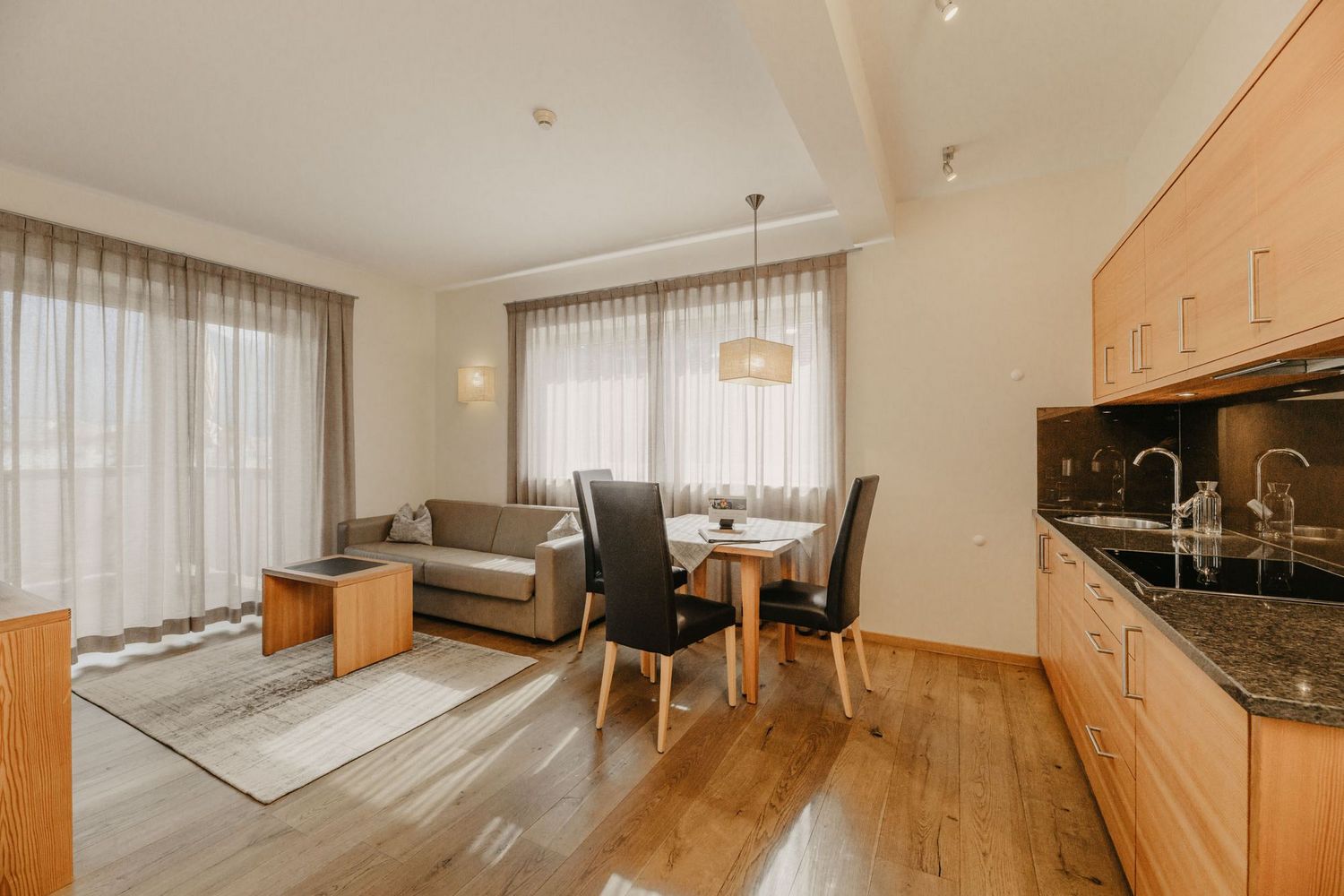 Ein offener Wohnbereich mit einer Kochnische mit Holzfronten, einem Sofa, einem kleinen Couchtisch und einem Essbereich mit zwei schwarzen Stühlen und einem Holztisch vor bodentiefen Fenstern mit hellen Vorhängen.