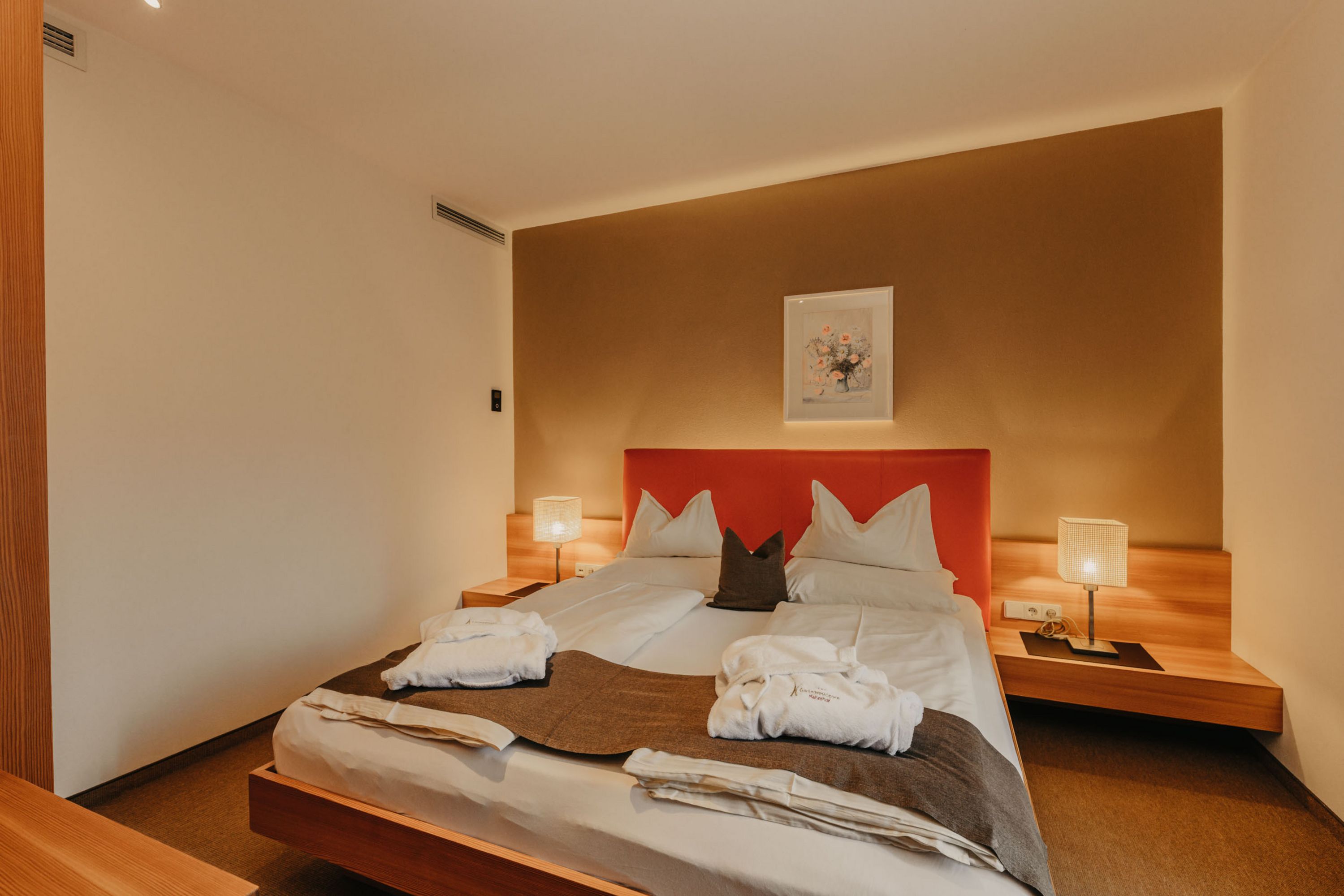Ein gemütliches Hoteldoppelzimmer mit einem großen Bett, das mit weißen Bettdecken, zwei Bademänteln und einem grauen Läufer ausgestattet ist, flankiert von zwei Nachttischlampen.