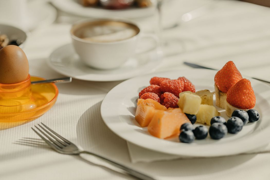 Ein Frühstückstisch mit einem gekochten Ei im orangen Eierbecher, einer Tasse Kaffee und einem Teller mit frischem Obstsalat aus Himbeeren, Blaubeeren, Ananas, Melone und Erdbeeren auf Bananenscheiben.