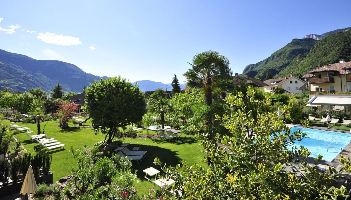 Un soleggiato giardino d'albergo con piscina, sedie a sdraio su un prato, palme e alberi davanti a uno scenario montano sotto un cielo azzurro.