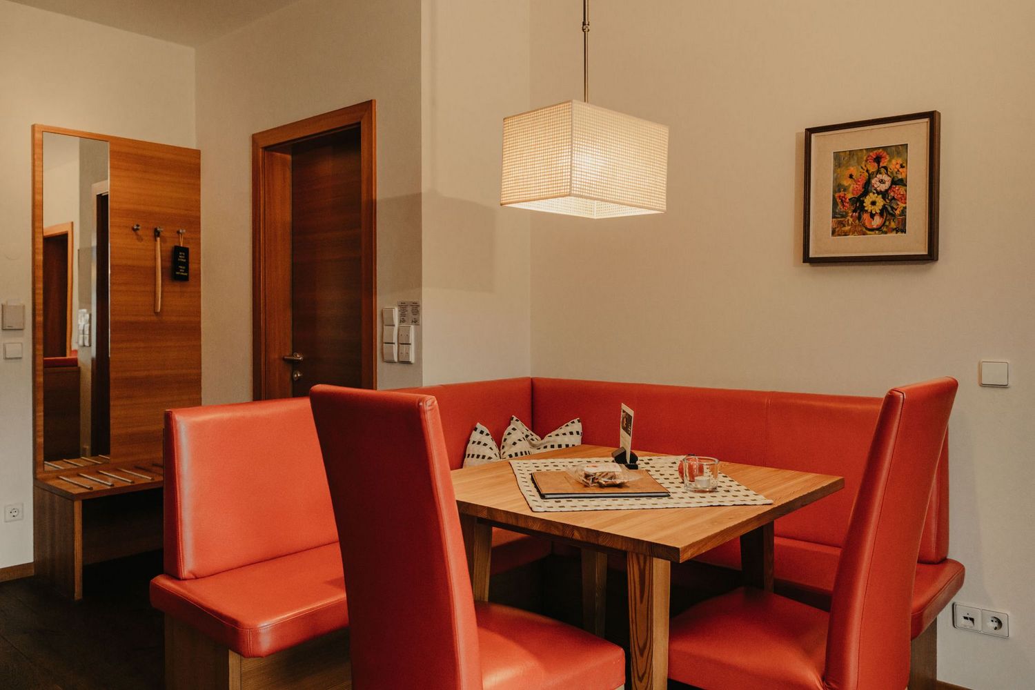 Eine gemütliche Sitzecke mit roter Eckbank und Holztisch, darüber eine moderne quaderförmige Deckenlampe in einem hellen Raum.