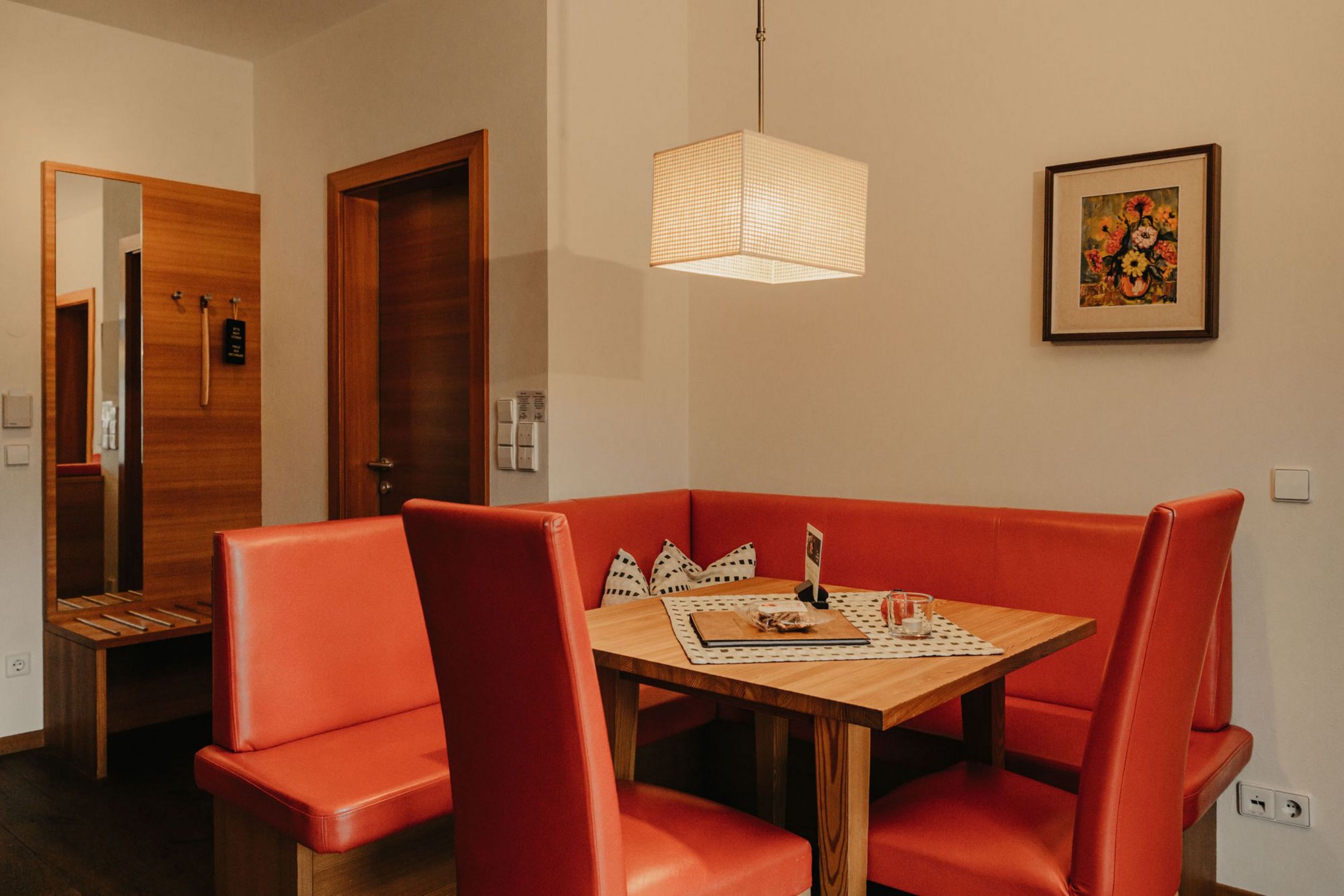 Eine gemütliche Sitzecke mit roter Eckbank und Holztisch, darüber eine moderne quaderförmige Deckenlampe in einem hellen Raum.