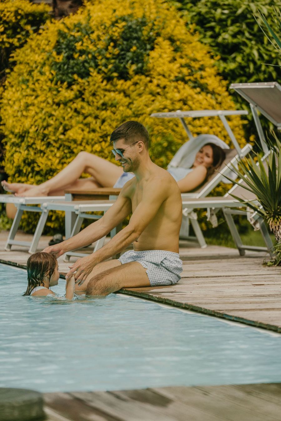 Ein Mann mit Badehose sitzt am Beckenrand eines Pools und spielt mit einem Kind im Wasser, während im Hintergrund eine Frau auf einer Sonnenliege entspannt.