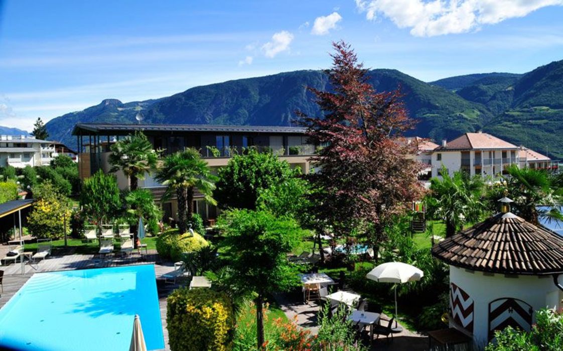 Ein sonniger Garten mit einem hellblauen Swimmingpool, umgeben von grünen Palmen, Sträuchern und Gebäuden vor einer imposanten Bergkulisse.