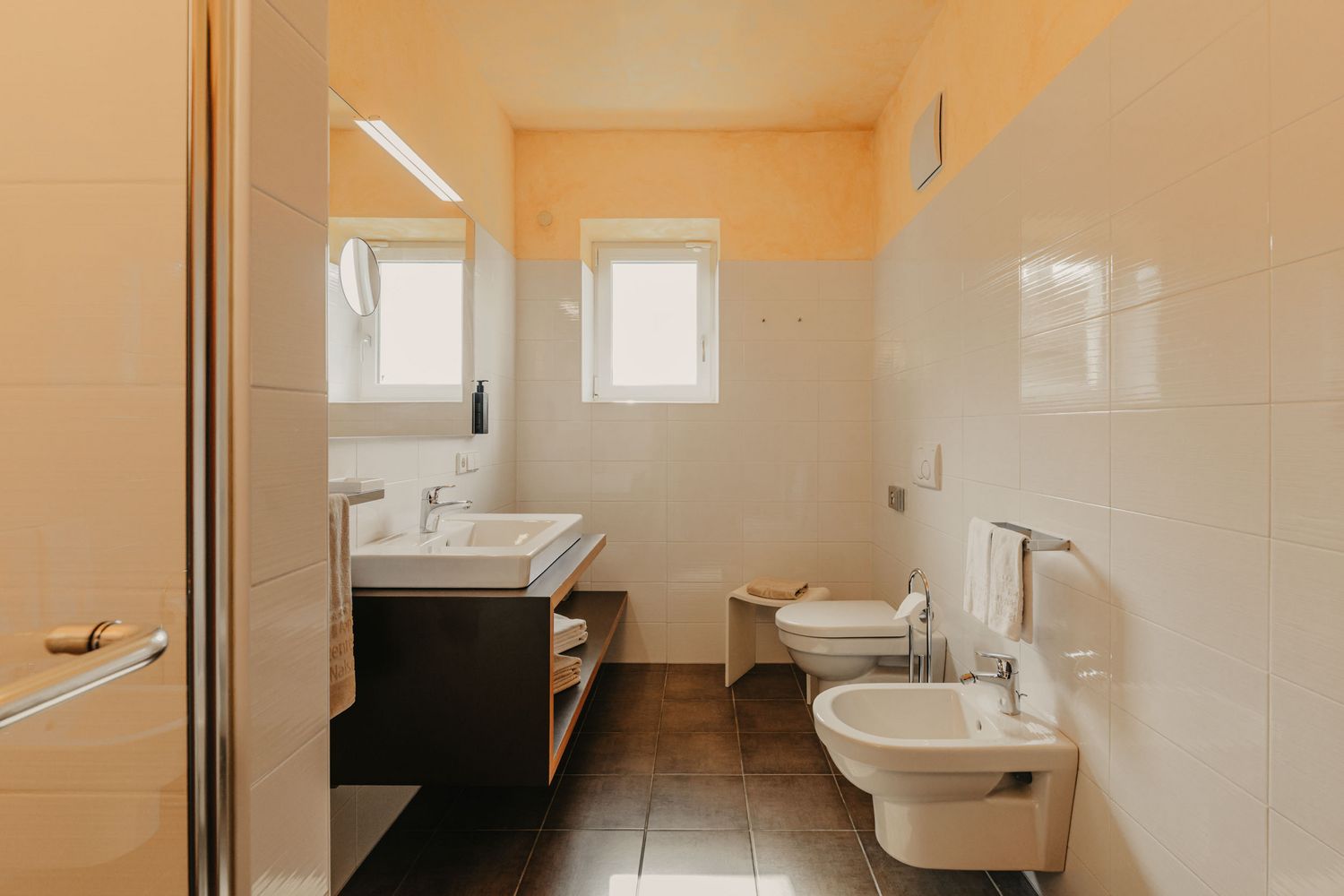 Ein Badezimmer mit dunkelbraunen Fliesen, weißgefliesten Wänden, einem Waschtisch mit Spiegel, einem WC und einem Bidet.
