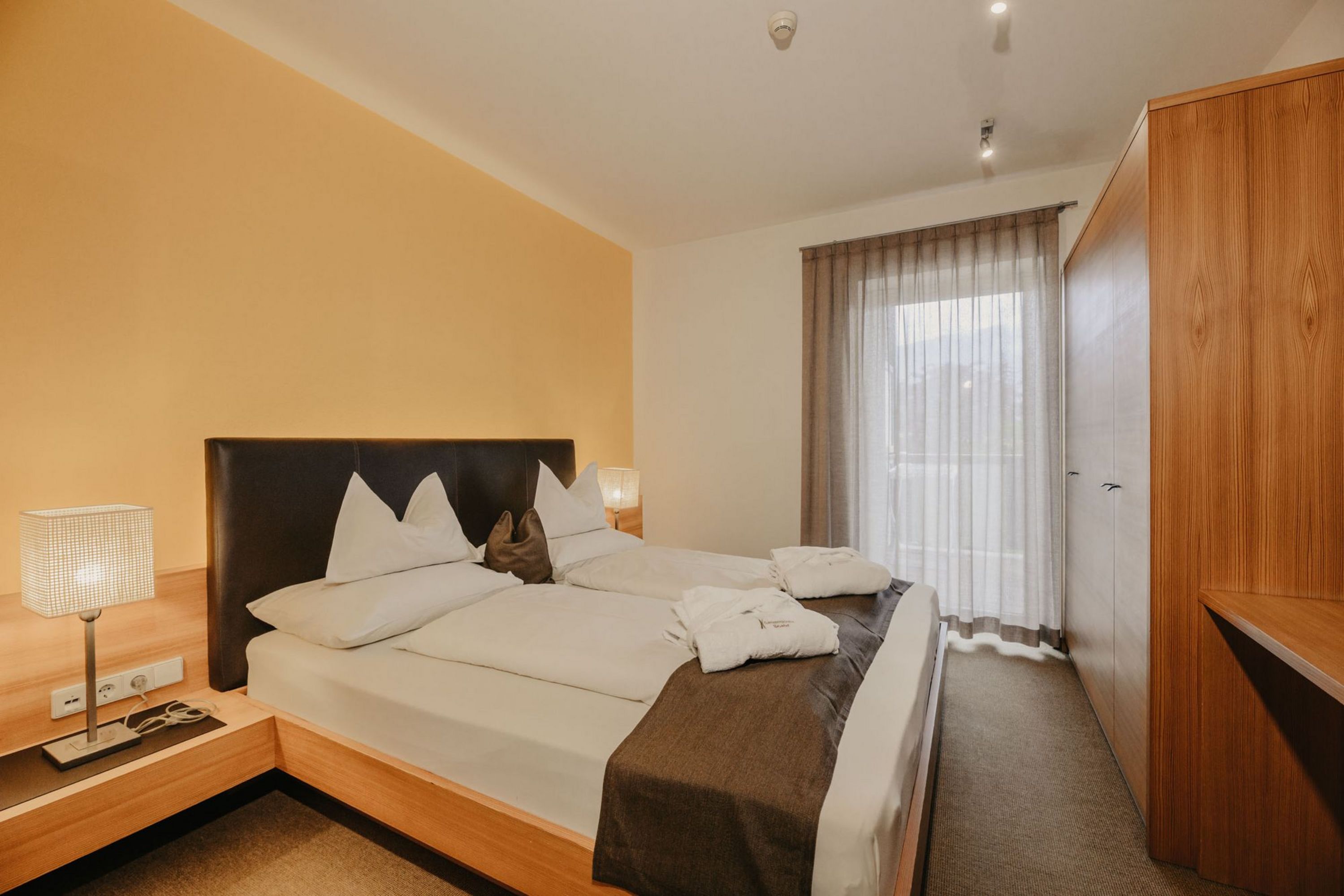 Ein ordentlich gemachtes Doppelbett mit weißer Bettwäsche und Bademänteln in einem Hotelzimmer mit Holzelementen und beigefarbener Wand.