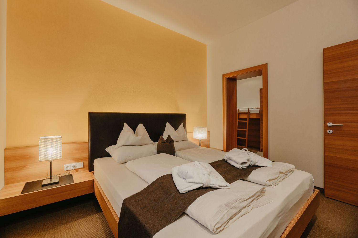 Ein ordentlich bezogenes Doppelbett mit zwei Nachttischlampen in einem Hotelzimmer mit gelber Wand und einem Durchgang zu einem weiteren Zimmer mit einem Hochbett.