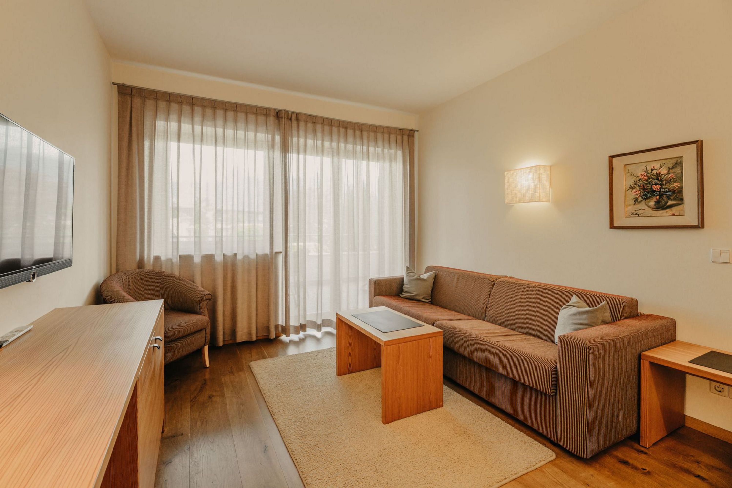 Ein gemütliches Hotelzimmer mit Sofa, Sessel, Couchtisch und einem Fernseher vor einem großen bodentiefen Fenster mit hellen Vorhängen.