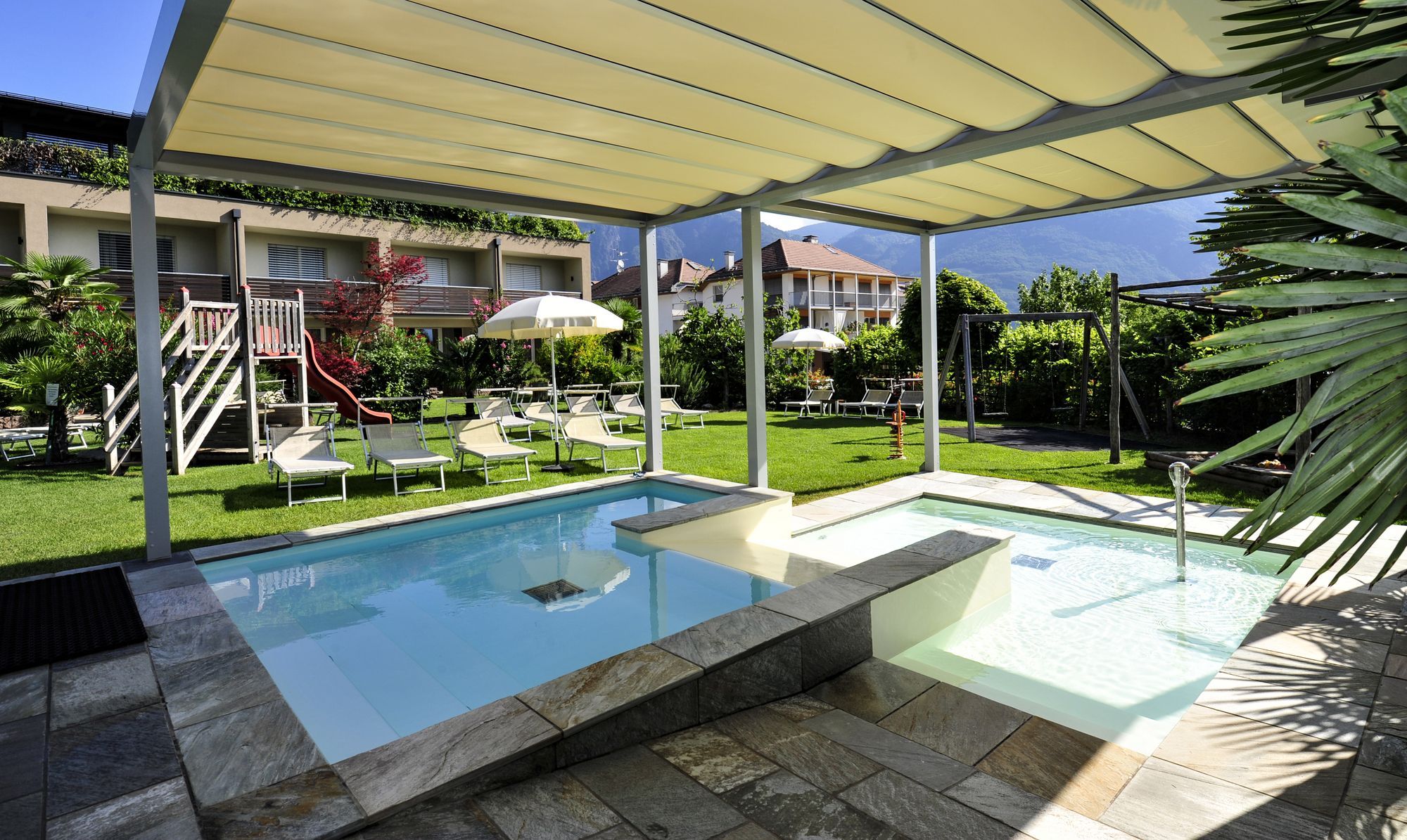 Ein Außenpool mit zwei Becken unter einer Pergola auf einem Hotelgelände mit Liegen, Sonnenschirmen und einem kleinen Spielplatz im Hintergrund.