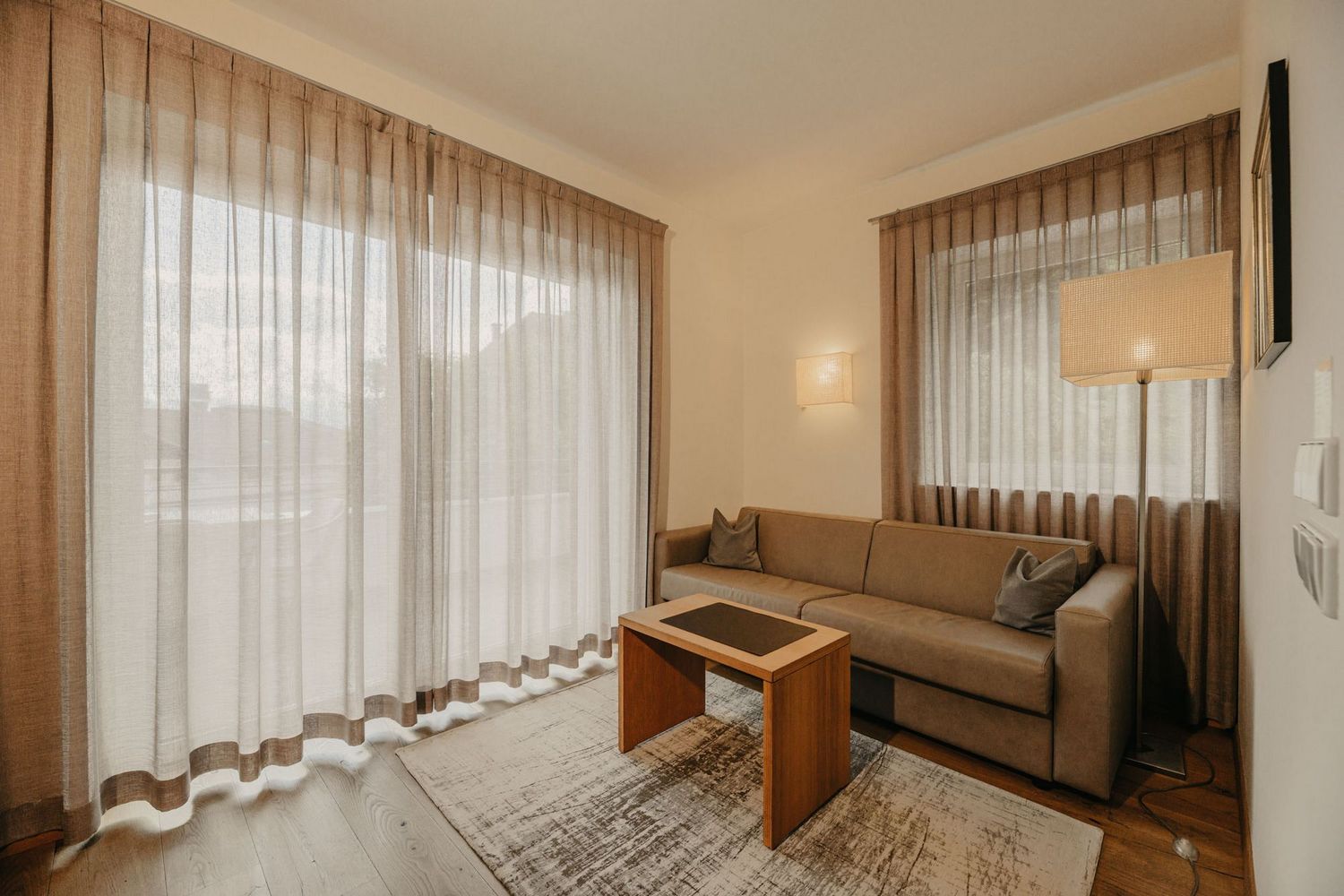 Ein gemütliches Wohnzimmer mit einem braunen Sofa, einem kleinen Holztisch, einem strukturierten Teppich und bodenlangen, beigen Vorhängen vor einem Fenster.