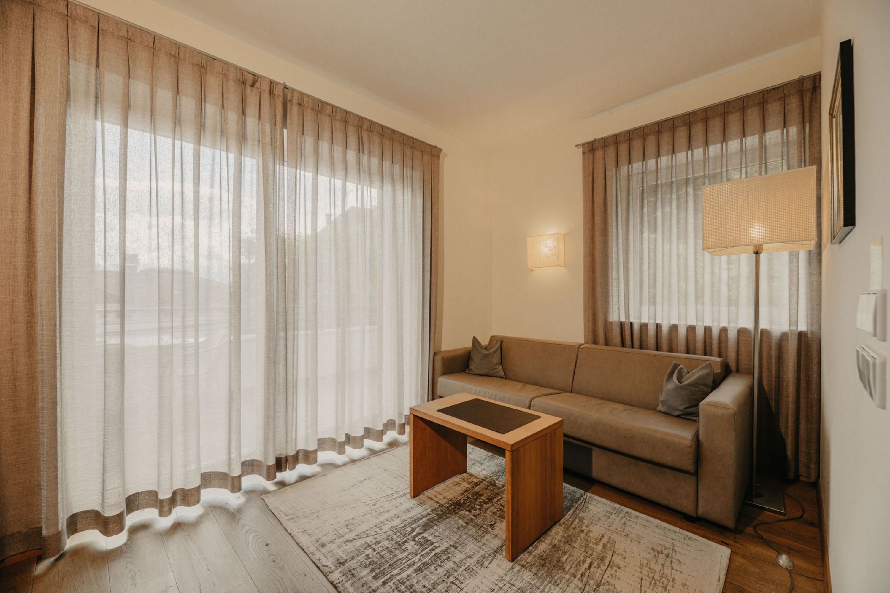 Ein gemütliches Wohnzimmer mit einem braunen Sofa, einem kleinen Holztisch, einem strukturierten Teppich und bodenlangen, beigen Vorhängen vor einem Fenster.