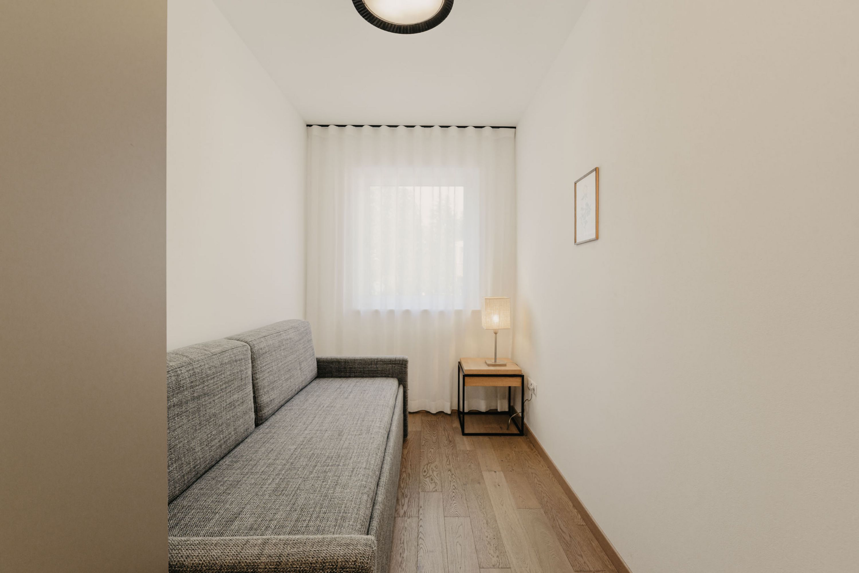 Ein schmales Gästezimmer mit hellen Wänden, Holzboden, einem grauen Schlafsofa und einem kleinen Beistelltisch mit einer Lampe vor einem Fenster mit weißem Vorhang.