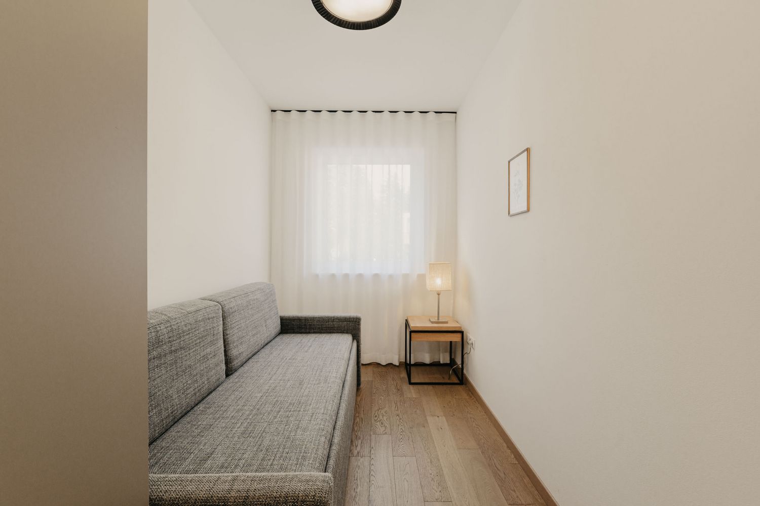 Ein schmales Gästezimmer mit hellen Wänden, Holzboden, einem grauen Schlafsofa und einem kleinen Beistelltisch mit einer Lampe vor einem Fenster mit weißem Vorhang.