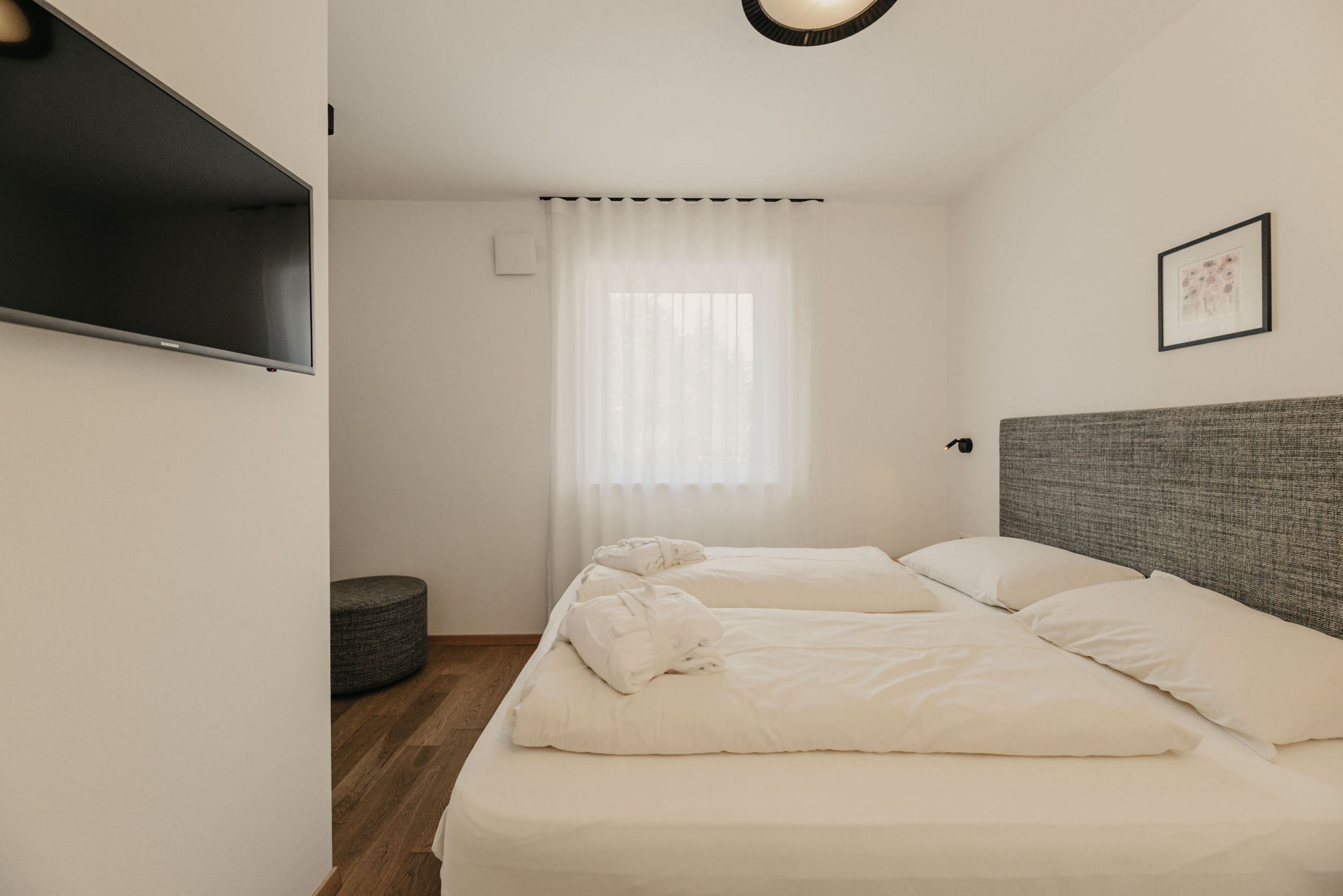 Ein minimalistisch eingerichtetes Hotelzimmer mit einem Doppelbett, zwei weißen Bademänteln, einem an der Wand montierten Fernseher und einem Fenster mit hellem Vorhang.