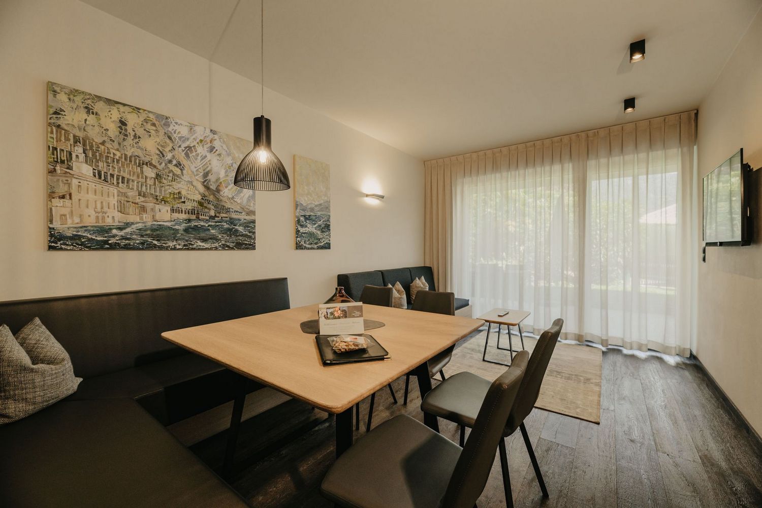 Ein modern eingerichtetes Wohn-Esszimmer mit einer dunklen Eckbankgruppe, Holztisch, Stühlen und einem Sofa vor einer großen Fensterfront mit hellem Vorhang.