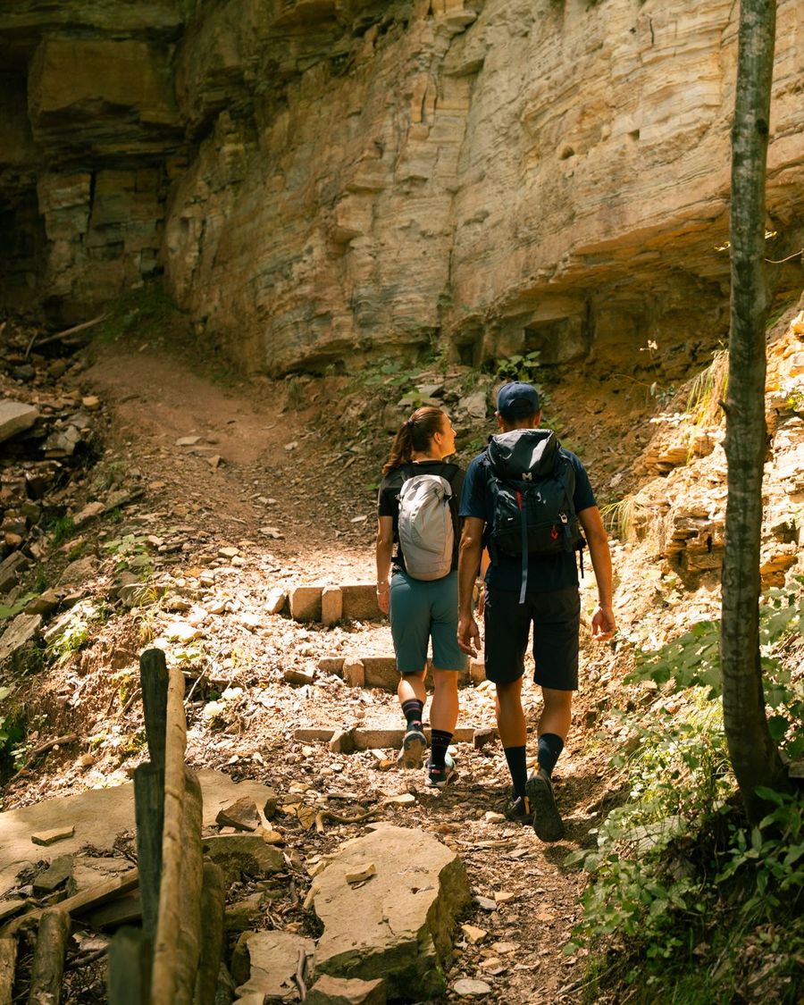 Ein junges Paar mit Wanderrucksäcken wandert auf einem schmalen, steinigen Pfad entlang einer steilen Felswand.