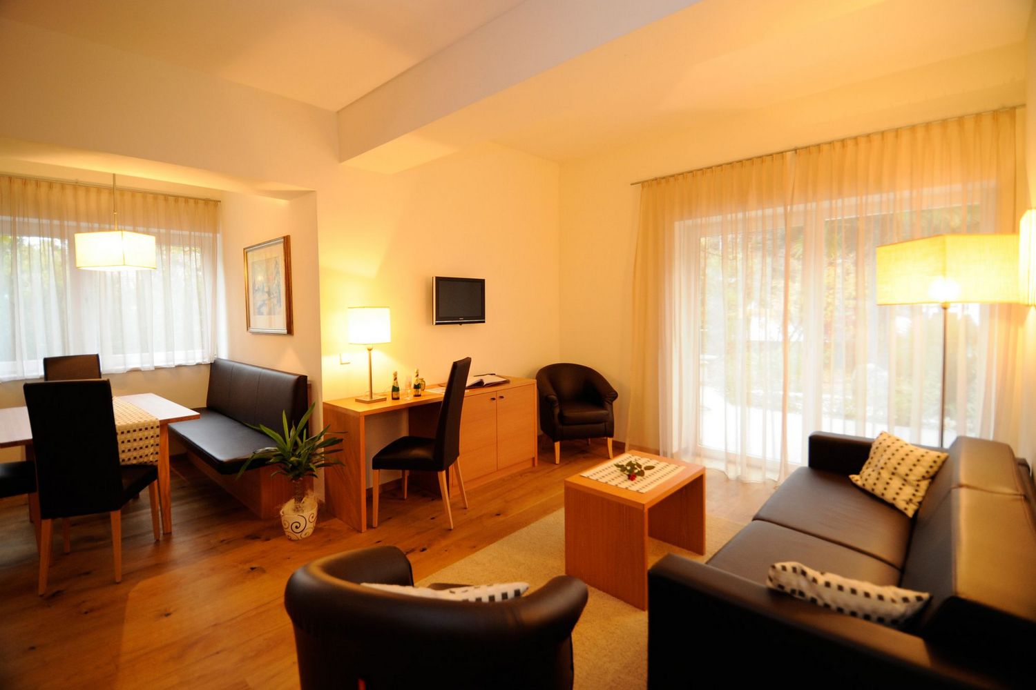 Ein gemütlicher Hotel-Wohnbereich mit Essplatz, Schreibtisch und einer Sitzecke mit schwarzem Sofa und Sesseln in warmem Licht.