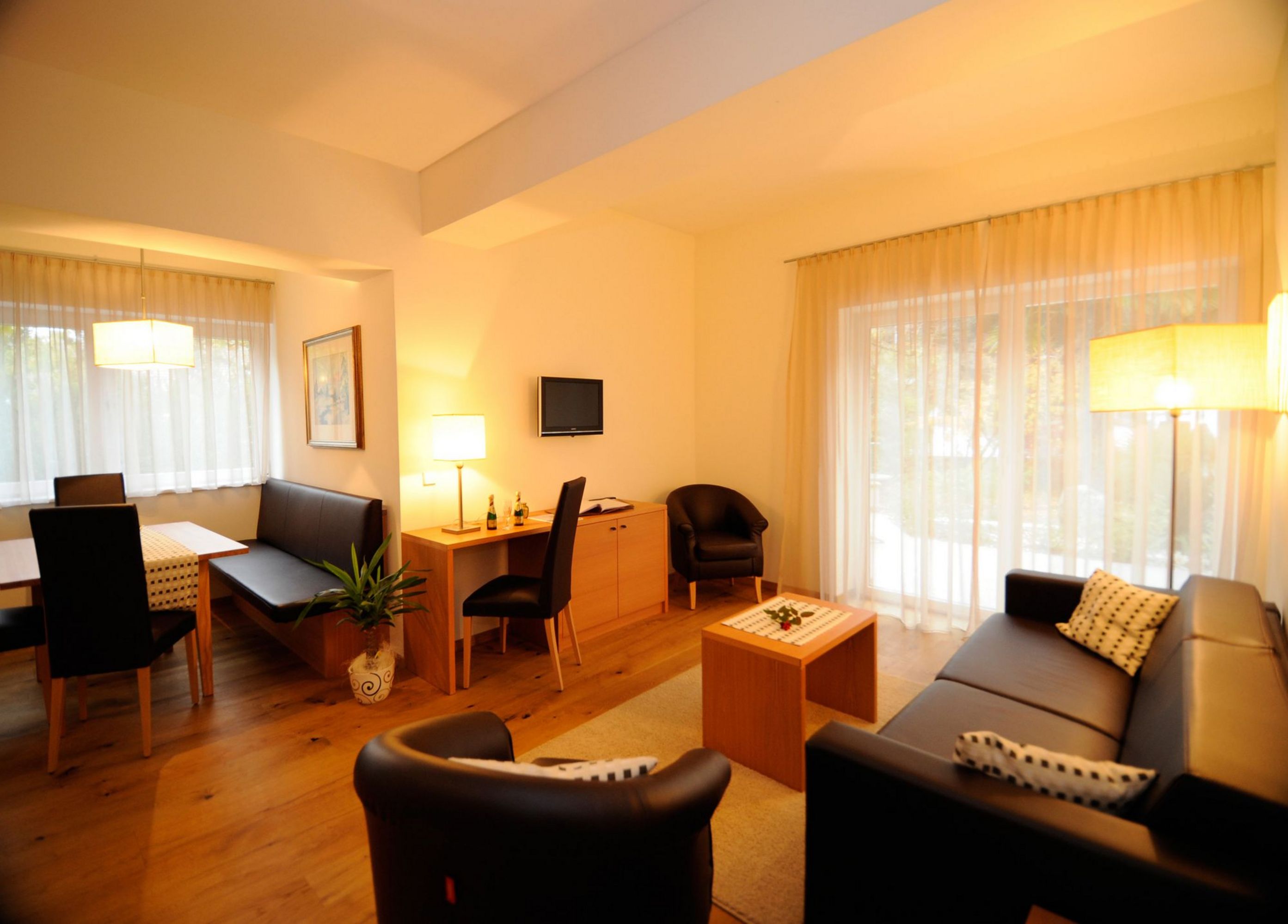 Ein gemütlicher Hotel-Wohnbereich mit Essplatz, Schreibtisch und einer Sitzecke mit schwarzem Sofa und Sesseln in warmem Licht.