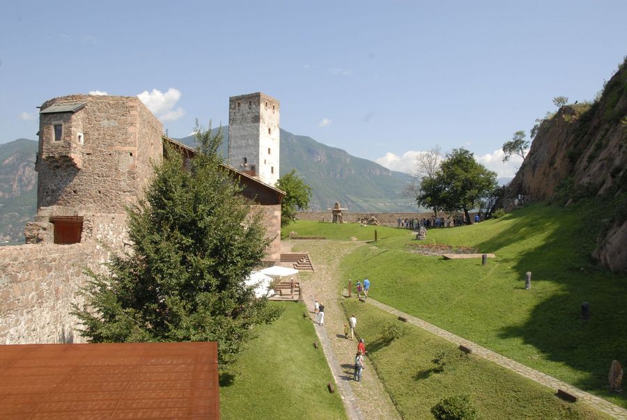 Ein sonniger Tag auf einer Burganlage mit alten Steinmauern, einem Wehrturm, einer grünen Wiese und Spaziergängern auf den Gehwegen.