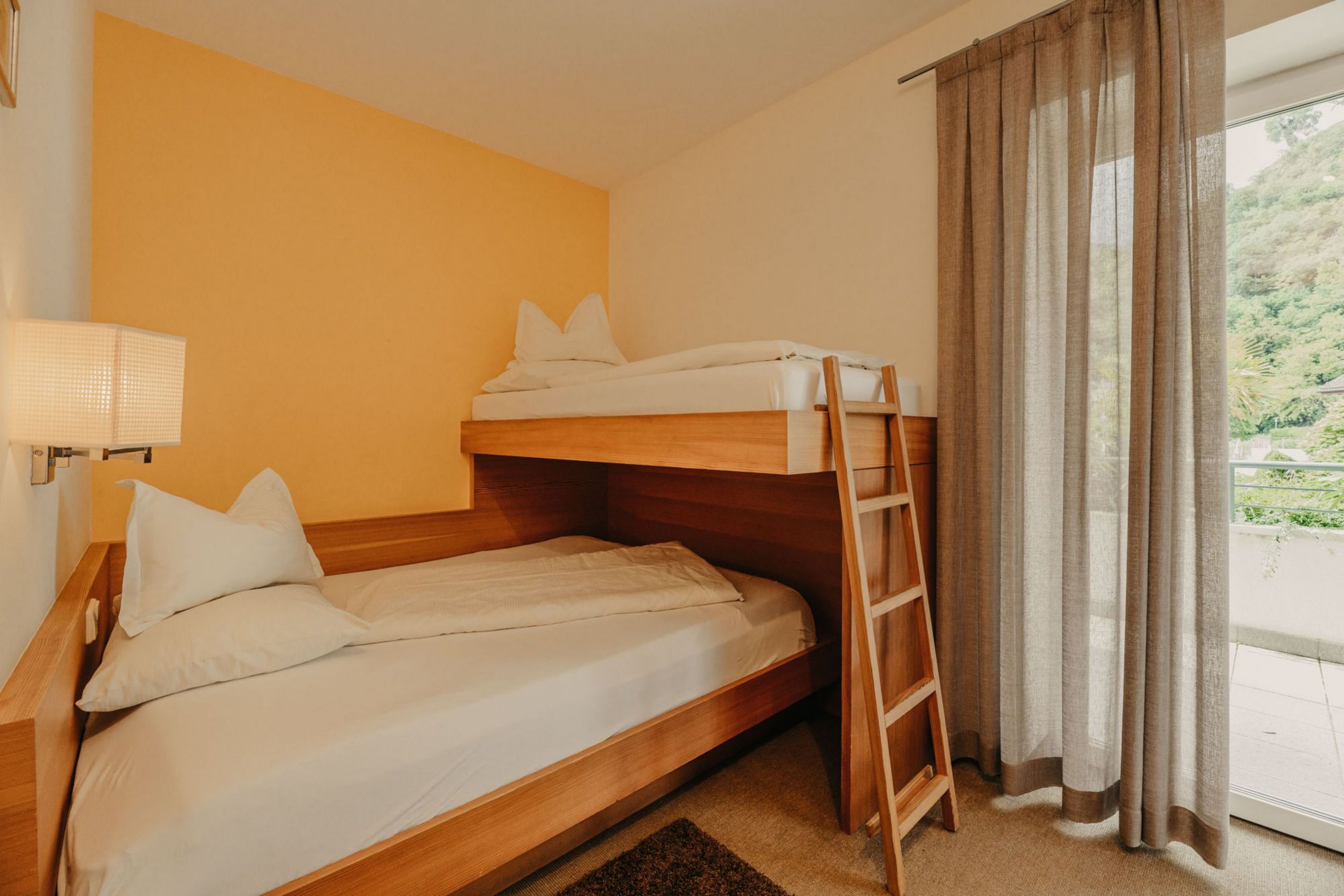 Ein gemütliches Hotelzimmer mit einem hölzernen Etagenbett, einer gelben Wand und einer Balkontür mit hellbraunen Vorhängen.
