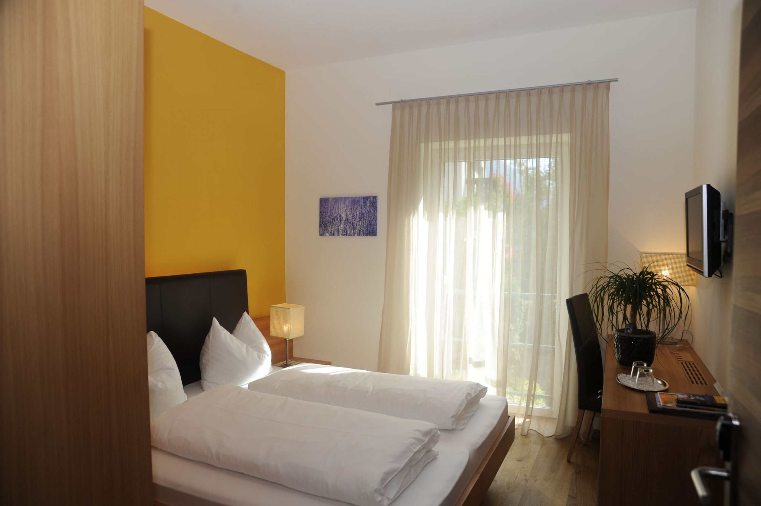 Ein helles Hotelzimmer mit einem Doppelbett, einer gelben Akzentwand, einem Schreibtisch mit Pflanze und einem bodentiefen Fenster mit hellen Vorhängen.