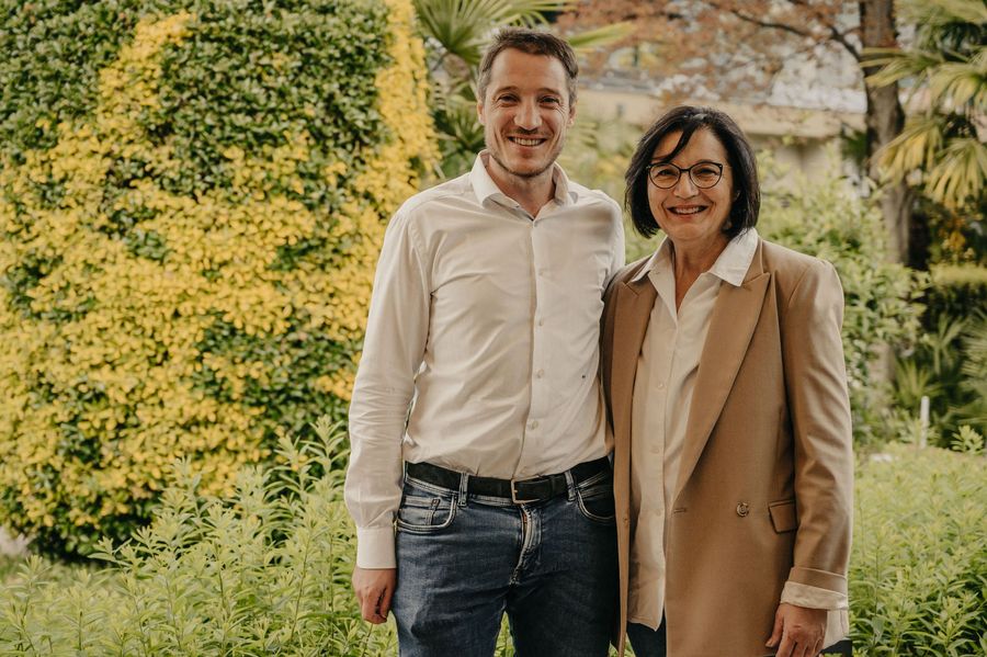 Ein Mann in einem weißen Hemd und eine Frau in einem sandfarbenen Blazer stehen nebeneinander im Freien vor einer begrünten Fläche.