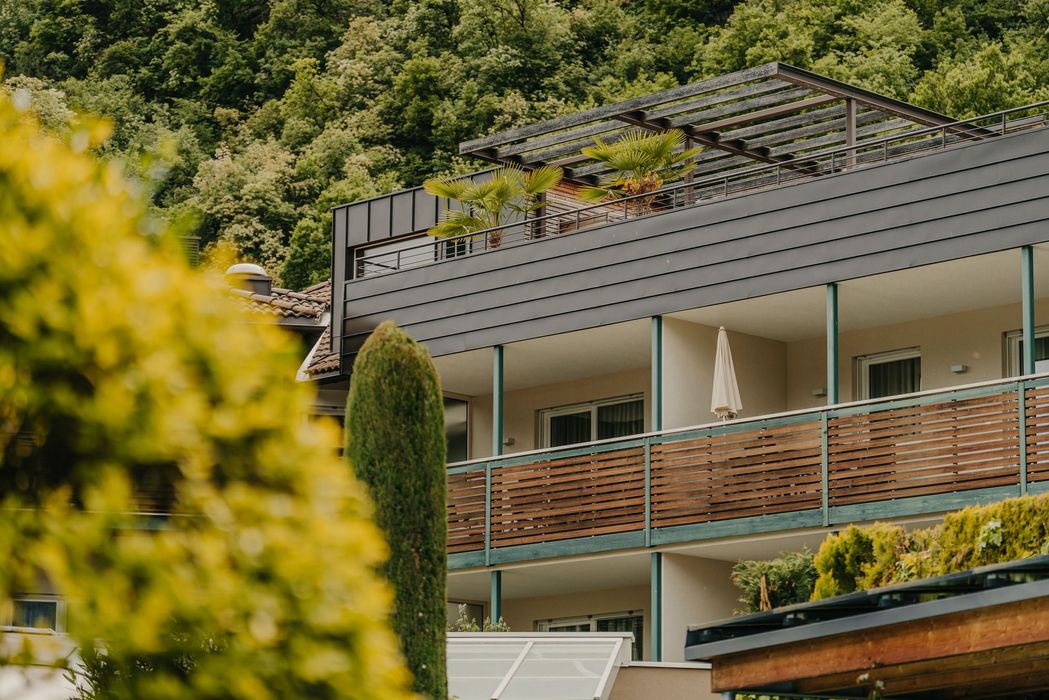 Ein modernes Gebäude mit einer dunkel verkleideten Dachterrasse, Holzbalkonen und Palmen vor einem bewaldeten Hang.